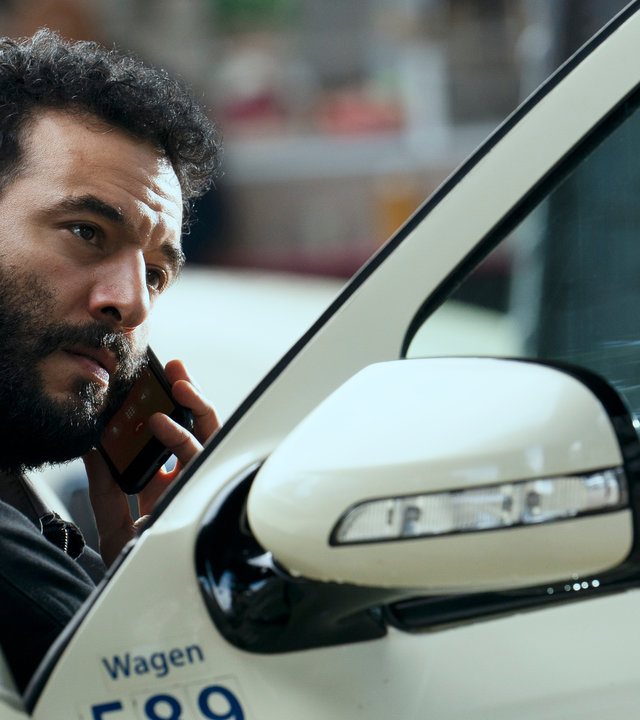 "LIBERAME - Nach dem Sturm - Wende": An einer geöffneten Autotür sitzt Ismail Sabia (Mohamed Achour). Mit der linken Hand, hält er sich ein Handy ans Ohr.