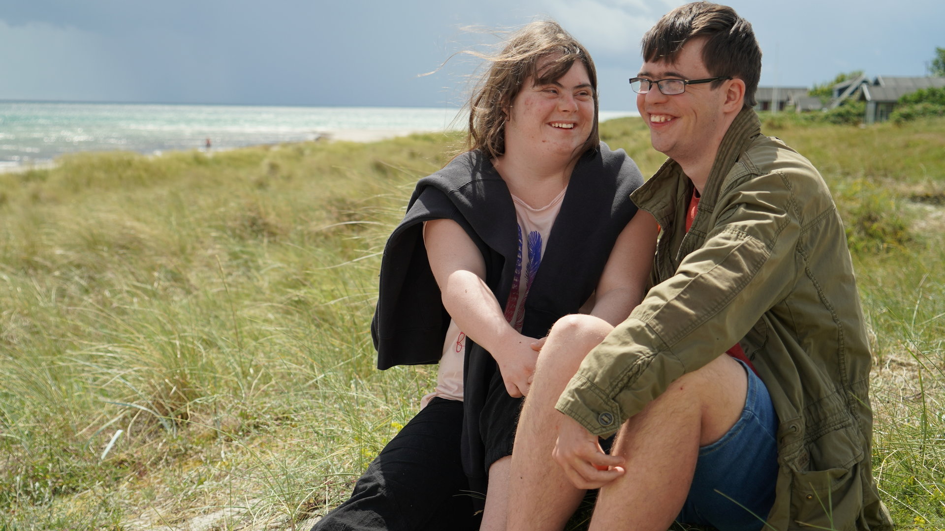 "37°: Liebe macht mutig - Zu Zweit mit Down Syndrom": Emily und Erik, jeweils mit Down-Syndrom, sitzen auf einer Düne am Strand und lächeln verliebt.