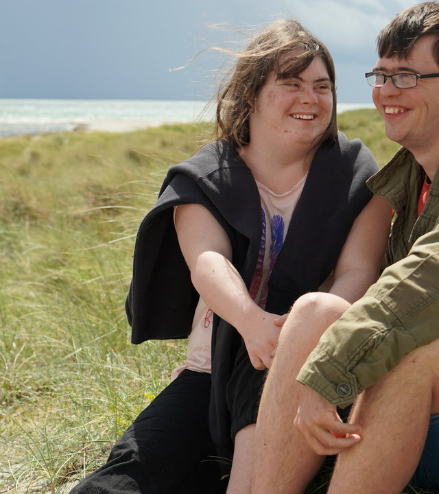 "37°: Liebe macht mutig - Zu Zweit mit Down Syndrom": Emily und Erik, jeweils mit Down-Syndrom, sitzen auf einer Düne am Strand und lächeln verliebt.