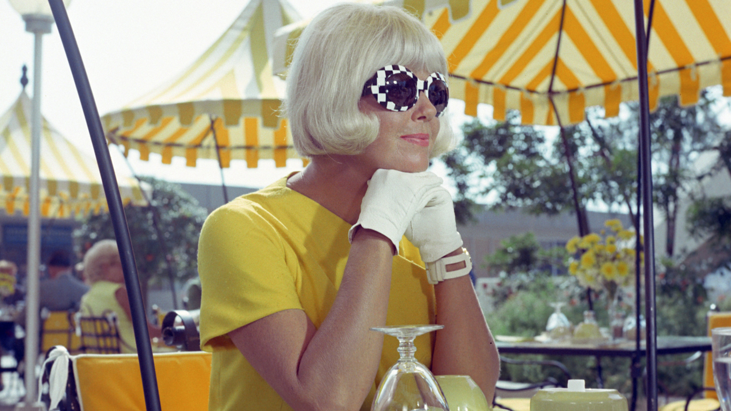 "Caprice": Patricia Fowler (Doris Day) sitzt im gelben Kurzarmkleid auf der Außenterrasse eines Lokals, ein Gedeck vor sich auf dem Tisch. Sie hat lächelnd ihr Kinn in ihren Händen abgestützt, die in weißen Handschuhen stecken. Ihre Augen sind verborgen hinter einer großen Sonnenbrille, deren Gestell ein exzentrisches, schachbrettartiges Muster zeigt.