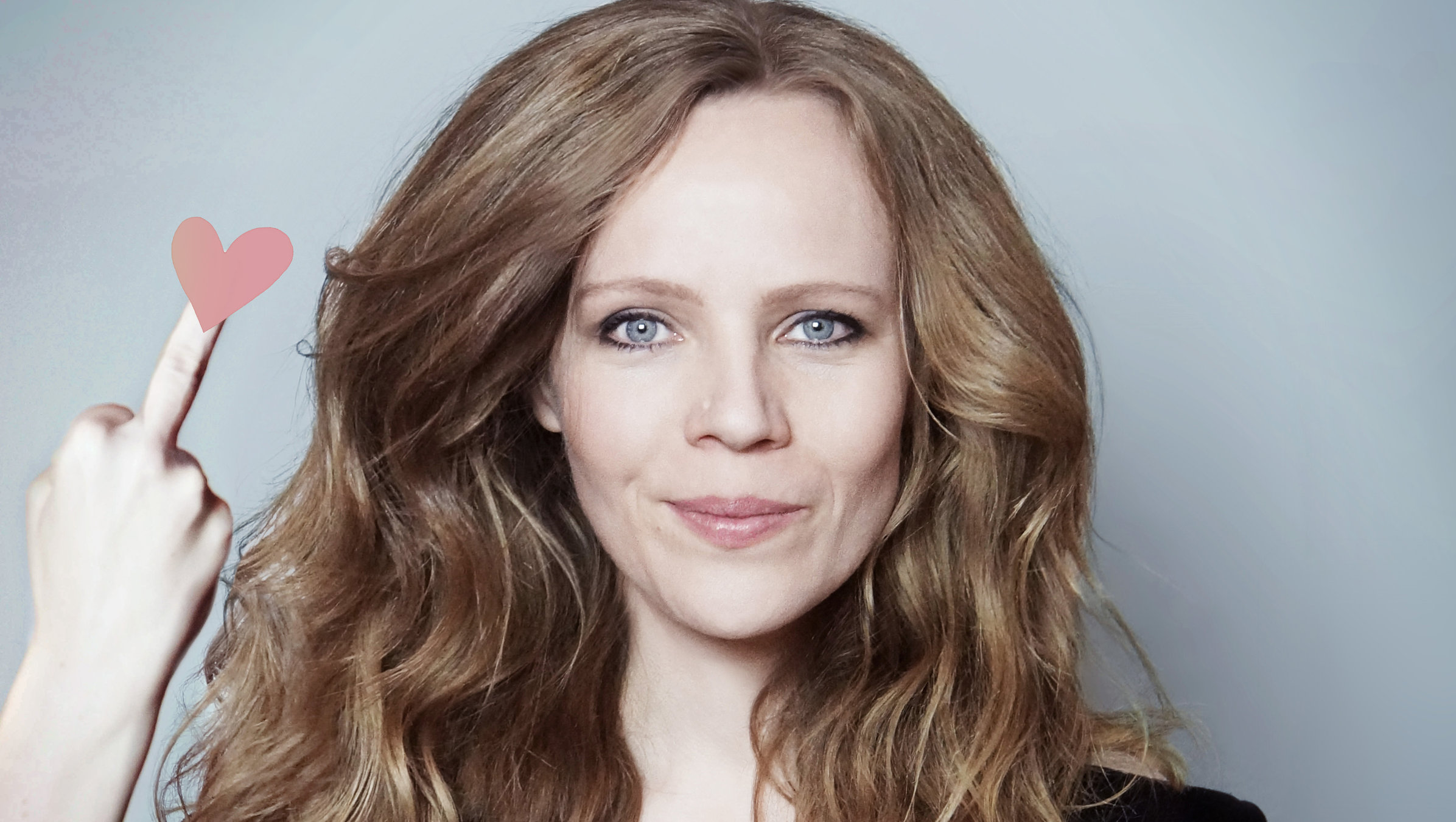 "Sarah Bosetti: Ich hab nichts gegen Frauen, du Schlampe!": Portrait von Sarah Bosetti.  Sie trägt ein schwarzes Oberteil und schaut freundlich in die Kamera, dabei zeigt sie ihren rechten Mittelfinger,  auf dem ein kleines rosa Herz ist.