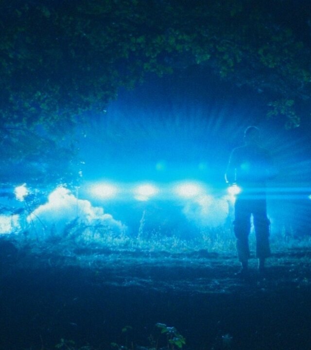 "Doppelhaushälfte – Ufo": Andi (Milan Peschel) steht in der Dunkelheit im Wald als Silhouette vor einer Reihe heller, blauer Lichter, die den Boden ausleuchten.