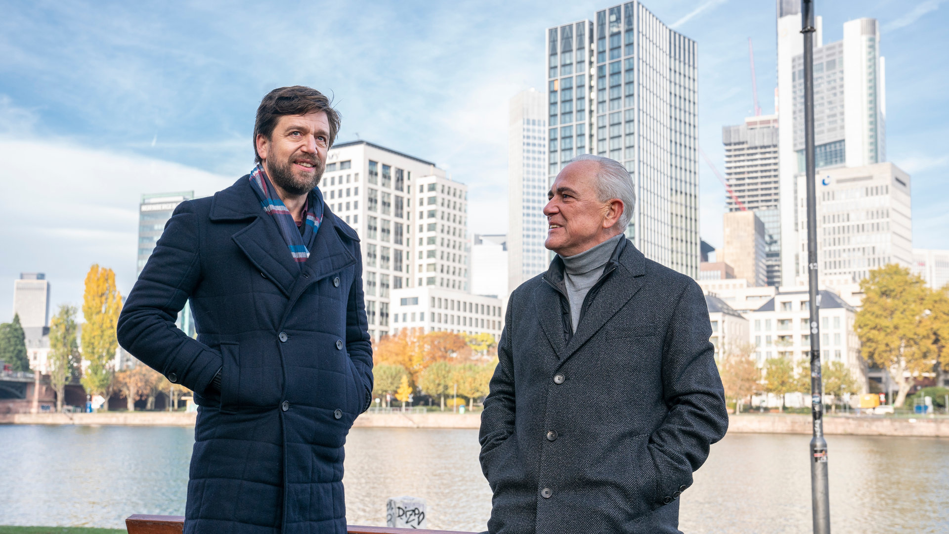 "The Bank Hacker": Alidor Van Praet (Gene Bervoets) lehnt sich an eine Parkbank und unterhält sich mit Bart (Koen De Graeve). Im Hintergrund sind viele Wolkenkratzer.