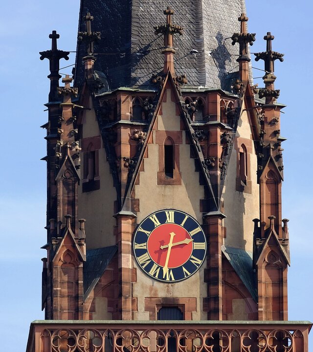 "Evangelischer Gottesdienst - Und Sara lachte": Auf dem Bild ist ein Teil des Turms der Dreikönigskirche in Frankfurt am Main zu seh. Filigrane, aus Stein gemeißelte Kreuze umrunden den Turm im neugotischen Stil, in seiner Mitte befindet sich die rote-blaue Kirchturmuhr mit goldenen Zeigern.