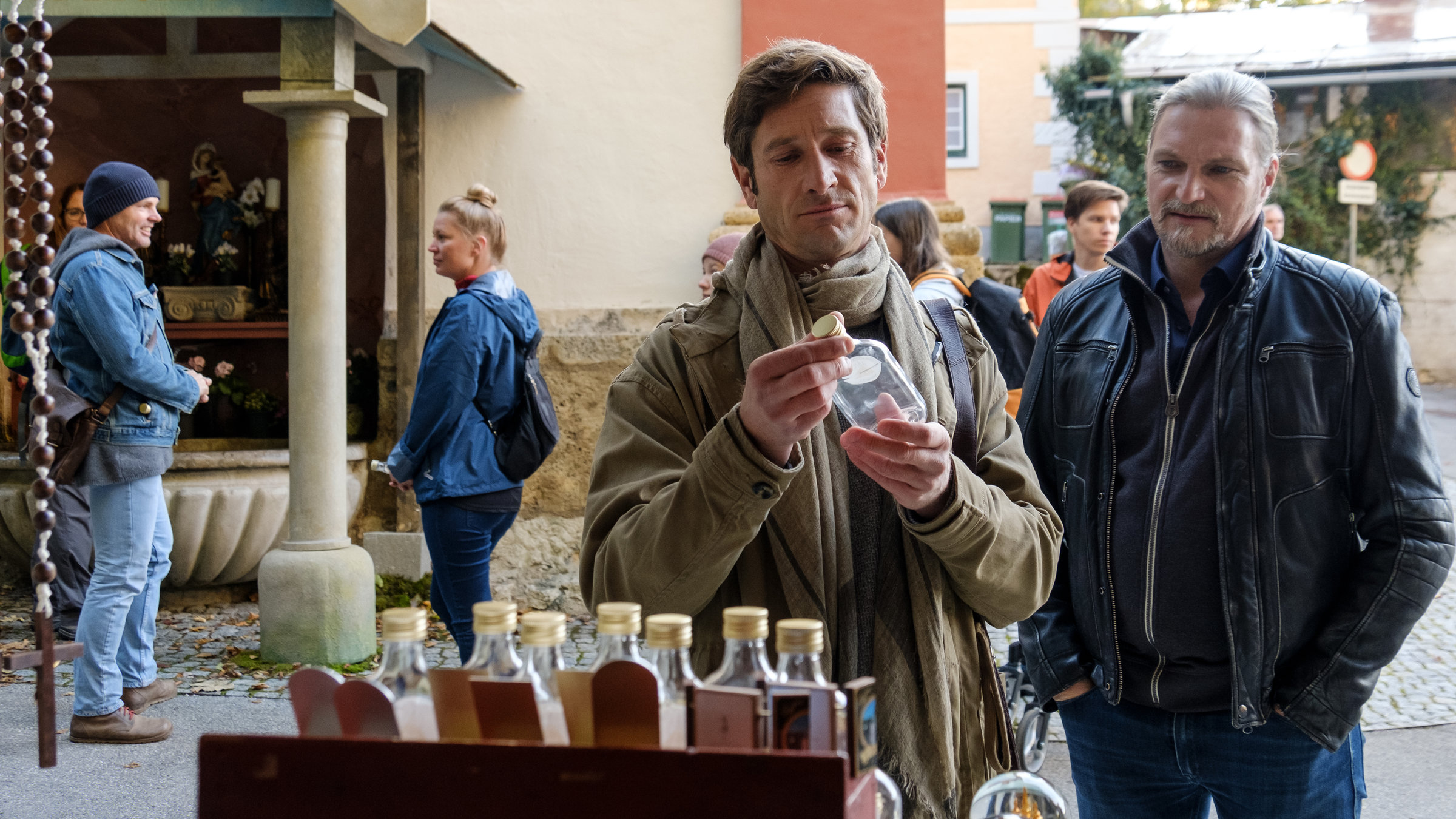 "SOKO Wien - Auf Abwegen": Bezirksinspektor Klaus Lechner (Andreas Kiendl) und Major Carl Ribarski (Stefan Jürgens) stehen vor einem Marktstand. Lechner halt eine kleine Glasflasche in den Händen und betrachtet sie eingehend. Carl Ribarski steht neben ihm und schaut zu.