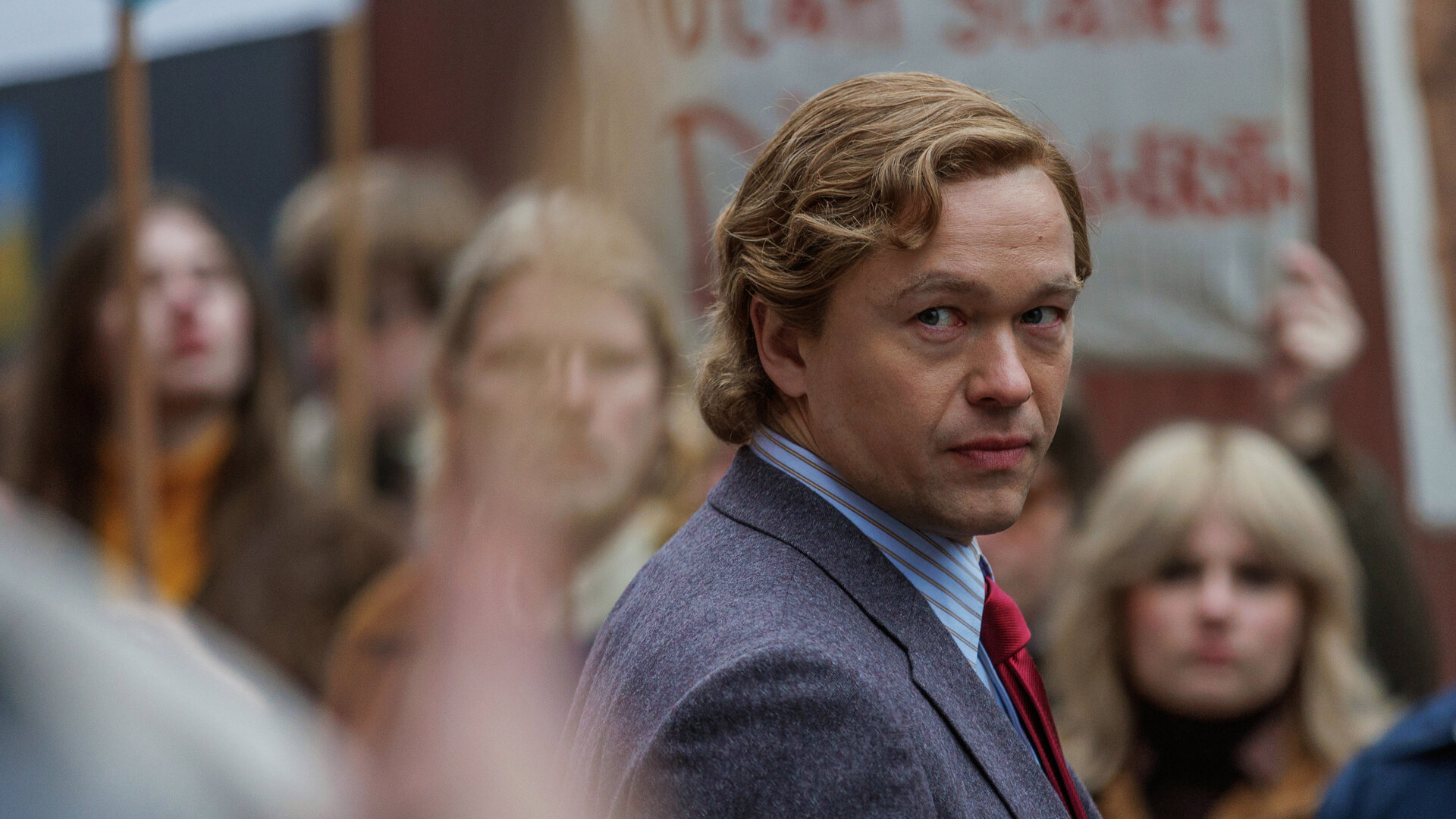 "Stenbeck - Der Aufstand des Erben": Jan Stenbeck (Jakob Oftebro) geht in Anzug auf einige Arbeiter (Komparsen) zu, die Protest-Plakate hochhalten, blickt jedoch von ihnen weg in Richtung Kamera.