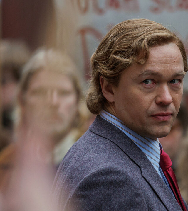 "Stenbeck - Der Aufstand des Erben": Jan Stenbeck (Jakob Oftebro) geht in Anzug auf einige Arbeiter (Komparsen) zu, die Protest-Plakate hochhalten, blickt jedoch von ihnen weg in Richtung Kamera.