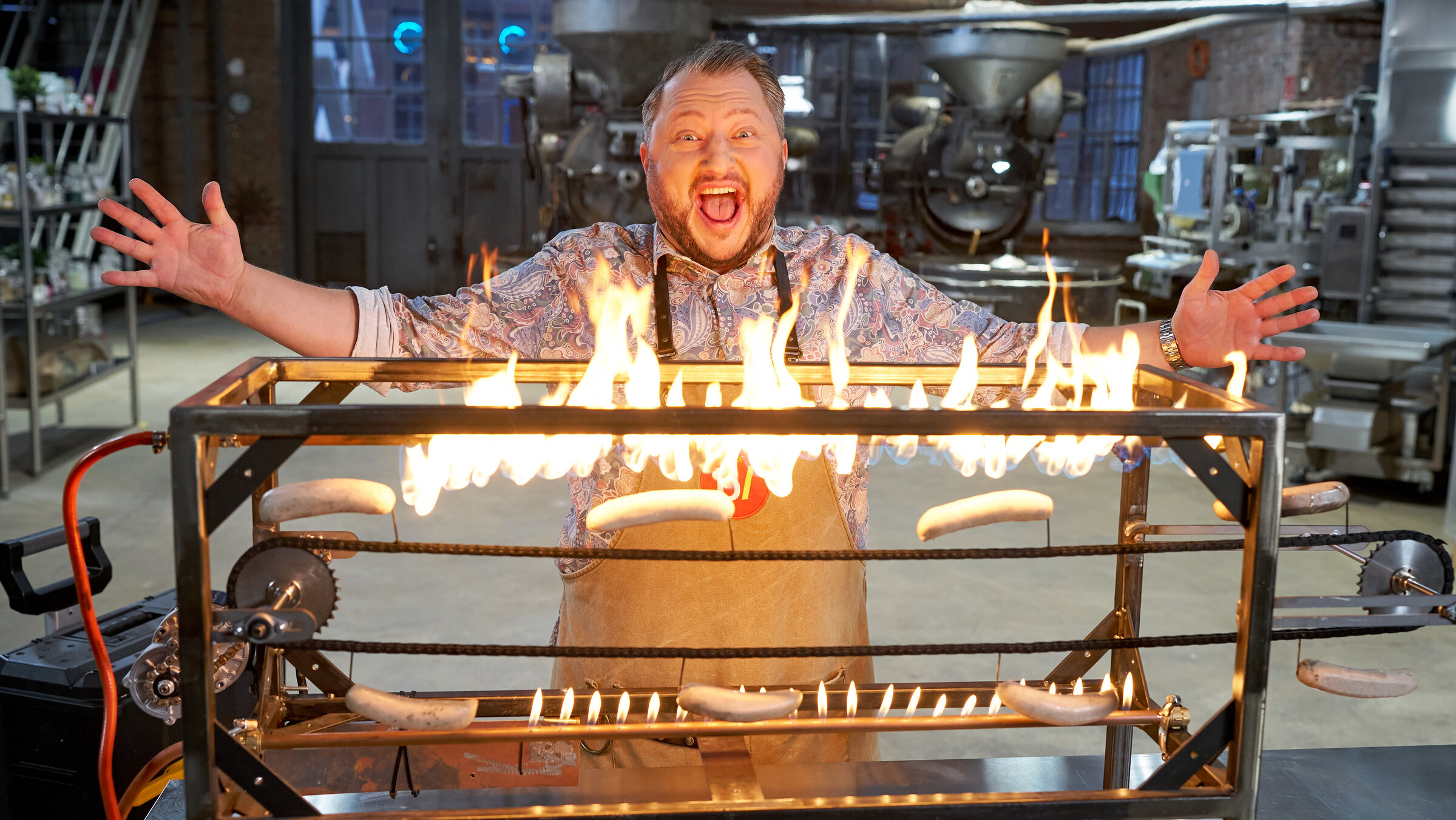 "besseresser: Die Tricks der Gastro-Industrie - Currywurst & Co.": Sebastian Lege steht mit ausgebreiteten Armen hinter einem selbst gebauten Würstchengrill, Flammen lodern auf und erhellen sein Gesicht.