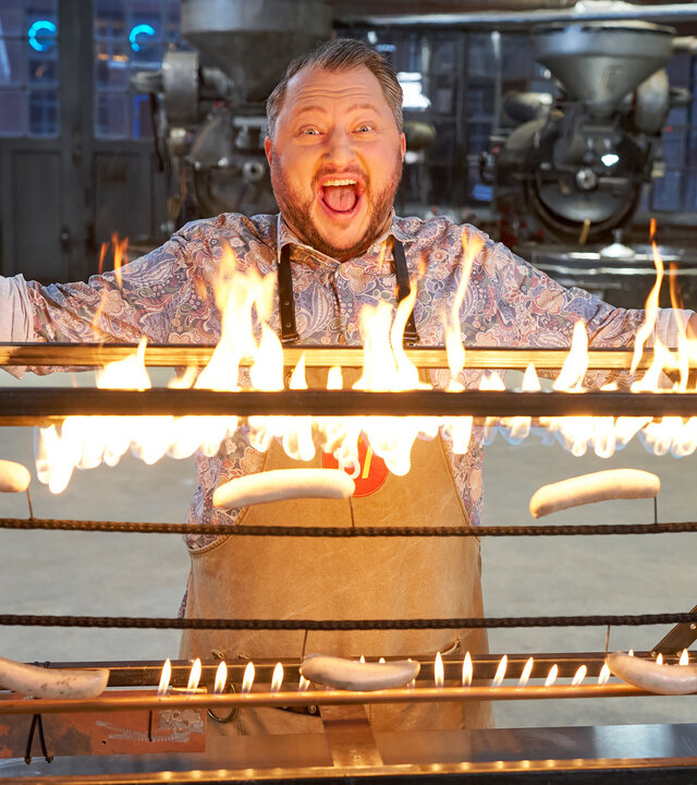 "besseresser: Die Tricks der Gastro-Industrie - Currywurst & Co.": Sebastian Lege steht mit ausgebreiteten Armen hinter einem selbst gebauten Würstchengrill, Flammen lodern auf und erhellen sein Gesicht.