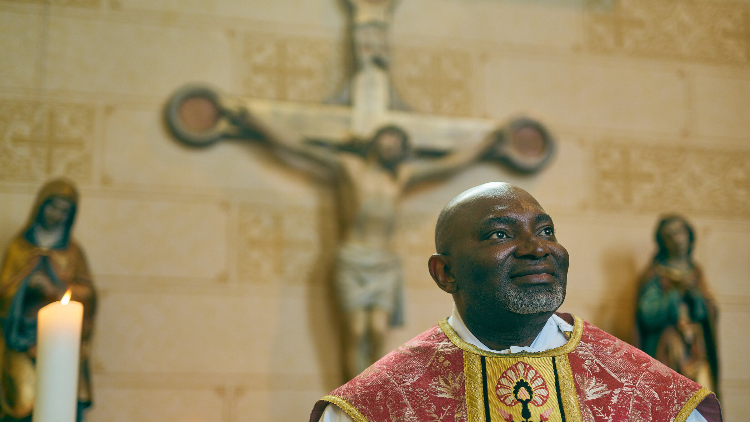"Kirche ohne Rassismus?": Der nigerianische Prieser Uchenna Aba steht vor einem Kreuz und zwei Figuren und schaut lächelnd nach oben. Neben ihm brennt eine Kerze.