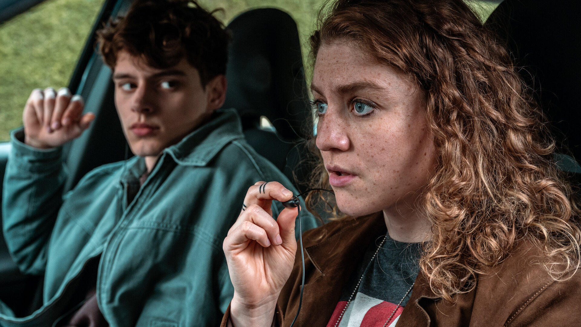 "Juliet - Roter Himmel": Juliet (Charlotte De Bruyne, r.) sitzt im Auto und spricht über ein kleines Mikrofon zwischen ihren Fingern mit Kollegen. Ihr junger Kollege Noah (Helder Onkelinx, l.) schaut ihr vom Beifahrersitz aus zu.