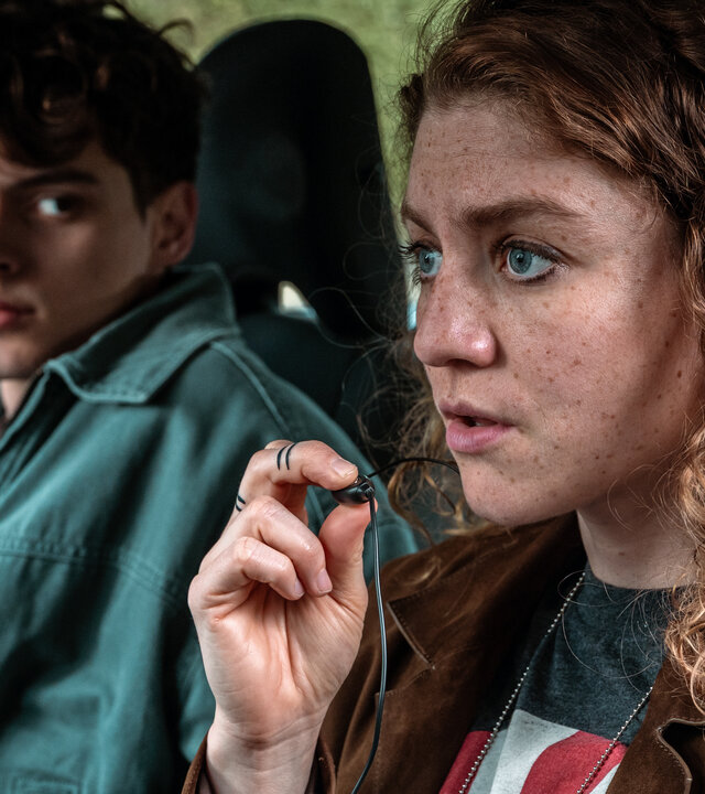 "Juliet - Roter Himmel": Juliet (Charlotte De Bruyne, r.) sitzt im Auto und spricht über ein kleines Mikrofon zwischen ihren Fingern mit Kollegen. Ihr junger Kollege Noah (Helder Onkelinx, l.) schaut ihr vom Beifahrersitz aus zu.