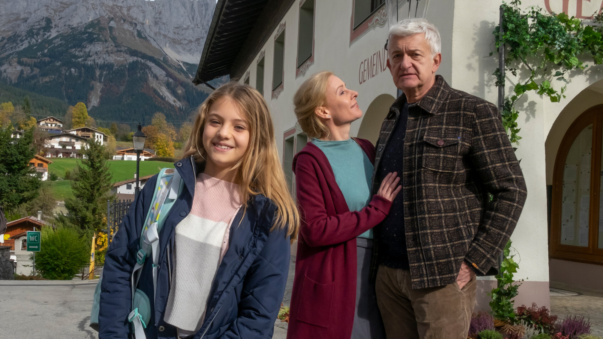 "Der Bergdoktor: Zerreißprobe (2)": Vor dem Gemeindehaus: Einen Rucksack über die Schulter tragend steht Sophie Gruber (Ylvi Unertl) lachend auf der Straße. Im Hintergrund stehen Susanne Dreiseitl (Natalie O'Hara) und Paul Richter (Dominic Raacke) Arm in Arm.