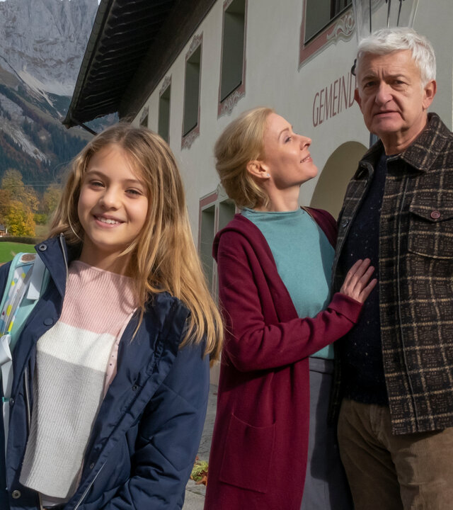 "Der Bergdoktor: Zerreißprobe (2)": Vor dem Gemeindehaus: Einen Rucksack über die Schulter tragend steht Sophie Gruber (Ylvi Unertl) lachend auf der Straße. Im Hintergrund stehen Susanne Dreiseitl (Natalie O'Hara) und Paul Richter (Dominic Raacke) Arm in Arm.