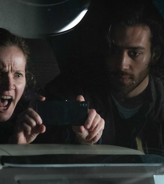 "Years and Years": Edith Lyons (Jessica Hynes) schreit, streckt dabei ihre Hände nach vorne und macht ein Foto. Neben ihr sitzt Viktor Goraya (Maxim Baldry) im Auto.