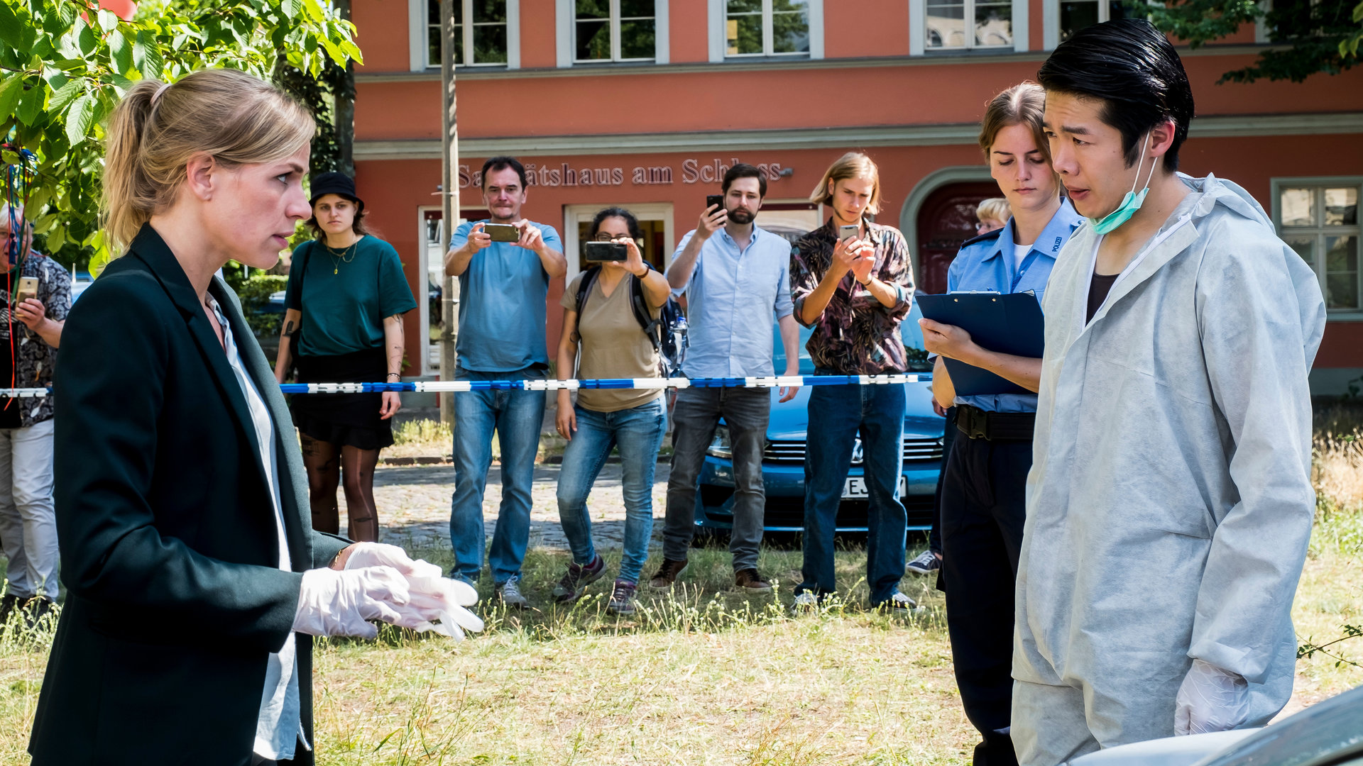 "SOKO Potsdam - Du bist schuld": Sophie Pohlmann (Katrin Jaehne) steht mit Thomas Brandner (Yung Ngo) auf einer Wiese. Thomas Brandner trägt einen weißen Schutzanzug und einen Mundschutz, den er nach unten gezogen hat. Sophie trägt weiße Gummihandschuhe. Die beiden blicken sich ernst an. Neben Thomas steht eine Polizistin (Komparsin) mit einem Klemmbrett in der Hand. Im Hintergrund stehen Passanten (Komparsen) hinter einem Absperrband und machen Fotos vom Tatort. Hinter ihnen sieht man ein Auto und ein Gebäude mit der Aufschrift "Sanitätshaus am Schloss". Die Sonne scheint.