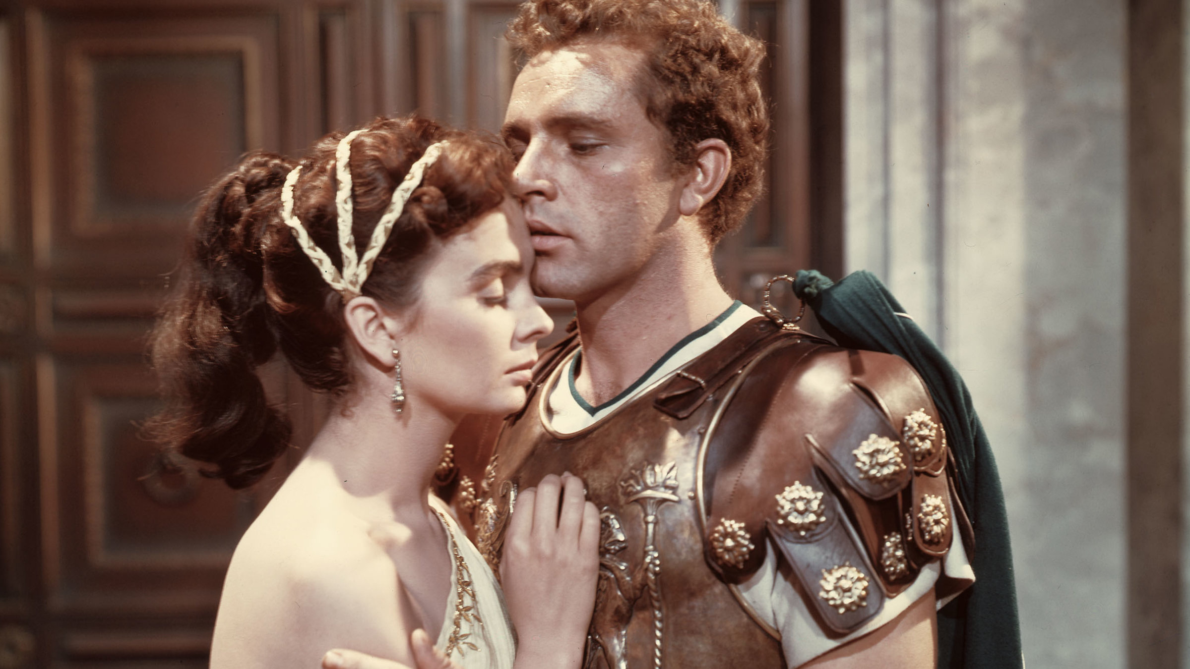 "Das Gewand": Jean Simmons (Diana, links) trägt eine weiße Tunika mit Gürtel und Kopfschmuck. Richard Burton (Marcellus, rechts) hat eine Panzerrüstung an. Das Paar umarmt sich liebevoll.