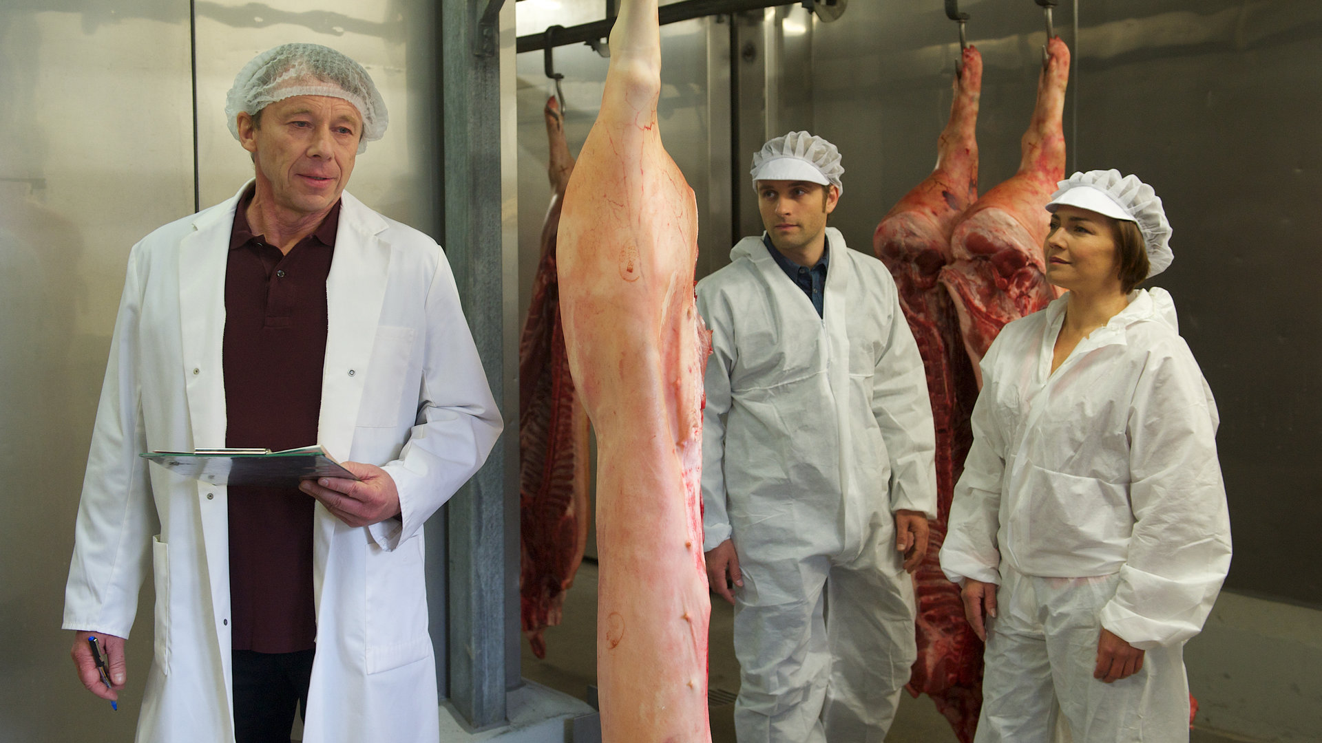 "SOKO Wismar - Erfroren": Nils Theede (Jonas Laux) und Katrin Börensen (Claudia Schmutzler), gekleidet in weiße Schutzanzüge, stehen in dem Kühlraum eines Schlachthofs und unterhalten sich mit dem Betriebsleiter Ole Grigor (Ulrich Wiggers). Zwischen ihnen hängen von der Decke frische Schweinehälften.