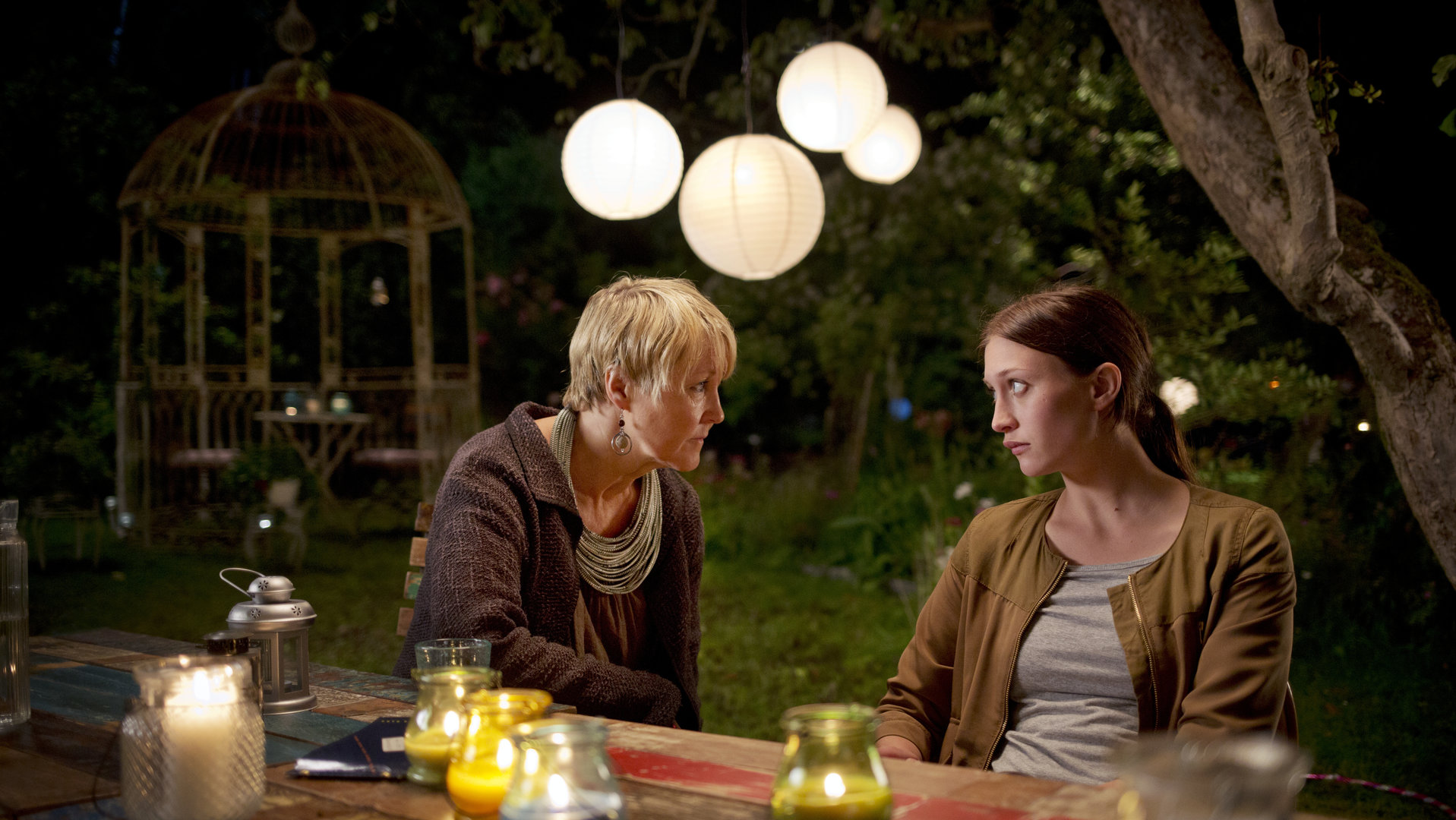 "Springflut - Erdrückender Beweis": Mette Olsäter (Cecilia Nilsson) und Olivia Rönning (Julia Ragnarsson) sitzen in Mettes Garten an einem Tisch und unterhalten sich. Vor ihnen stehen bunte, brennende Kerzen in Gläsern. Im Hintergrund hängen weiße runde Lampions am Baum.