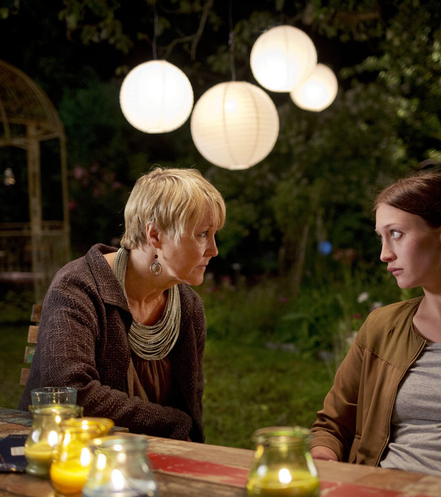 "Springflut - Erdrückender Beweis": Mette Olsäter (Cecilia Nilsson) und Olivia Rönning (Julia Ragnarsson) sitzen in Mettes Garten an einem Tisch und unterhalten sich. Vor ihnen stehen bunte, brennende Kerzen in Gläsern. Im Hintergrund hängen weiße runde Lampions am Baum.