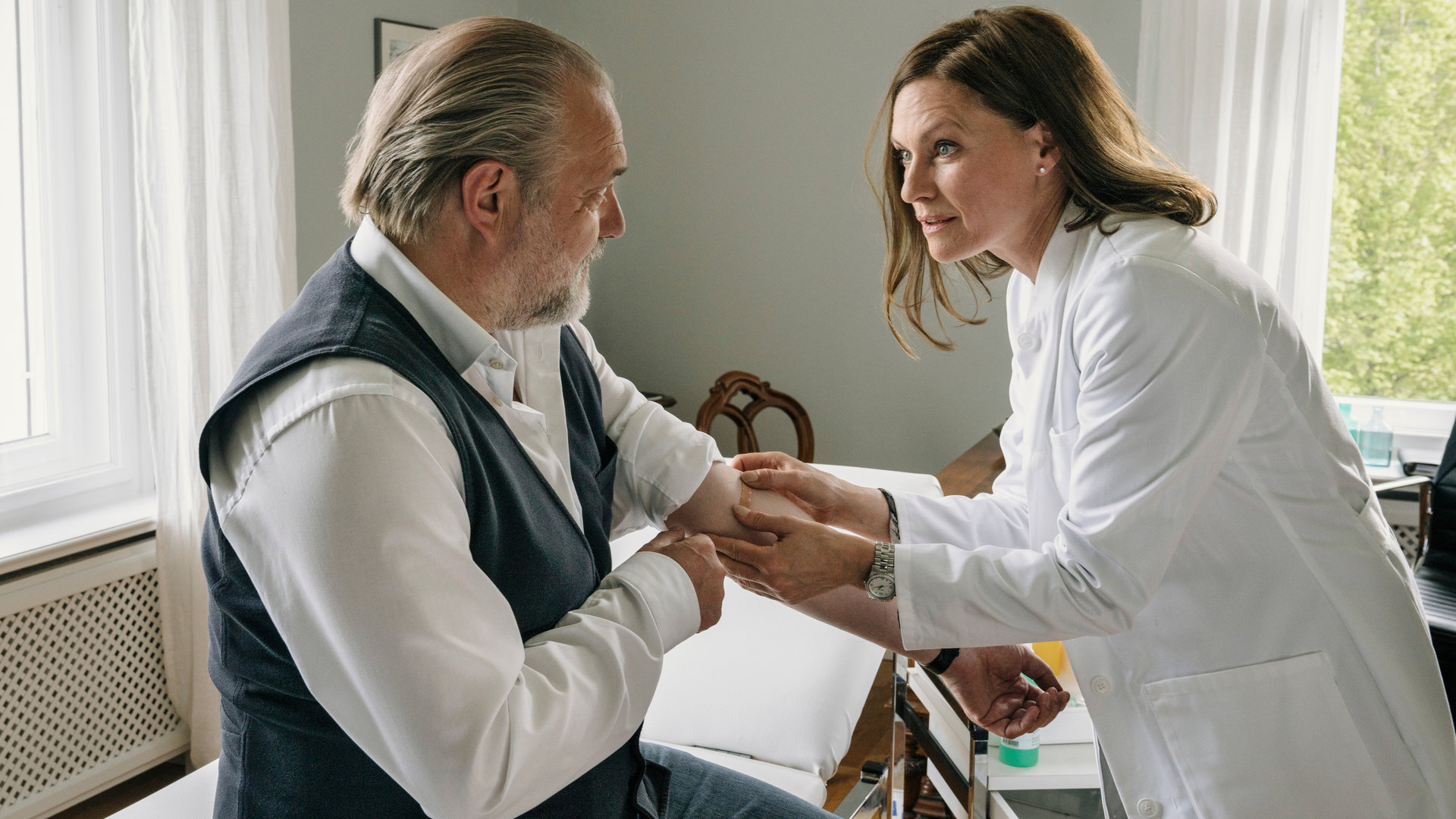 "Der Alte - Knock-out": Hauptkommissar Richard Voss (Jan-Gregor Kremp) sitzt in einem Behandlungszimmer auf einer Liege. Er hat linken Ärmel seines Hemds hochgekrempelt, damit Dr. Johanna Westhusen (Nina Kronjäger) ihm ein Pflaster aufkleben kann.