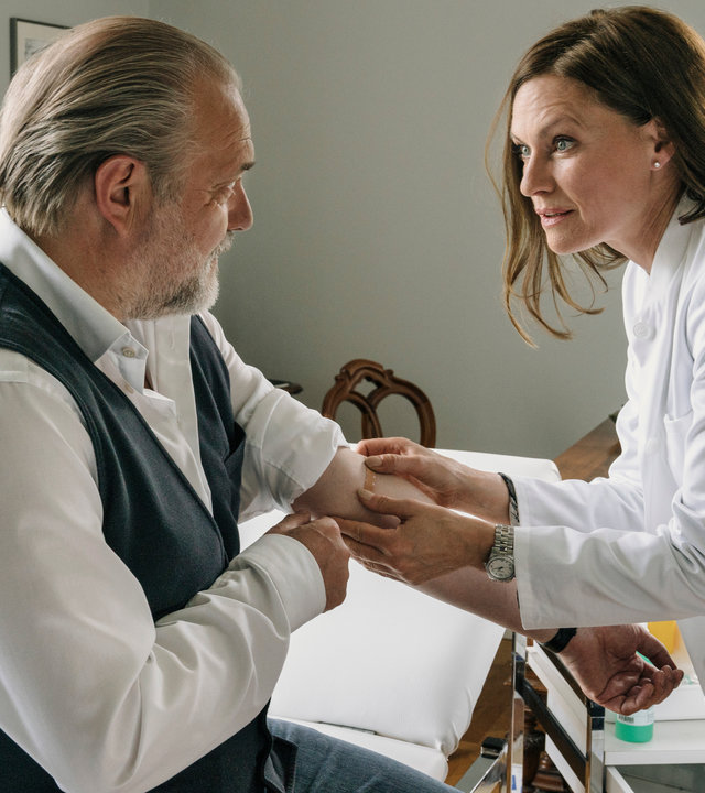 "Der Alte - Knock-out": Hauptkommissar Richard Voss (Jan-Gregor Kremp) sitzt in einem Behandlungszimmer auf einer Liege. Er hat linken Ärmel seines Hemds hochgekrempelt, damit Dr. Johanna Westhusen (Nina Kronjäger) ihm ein Pflaster aufkleben kann.
