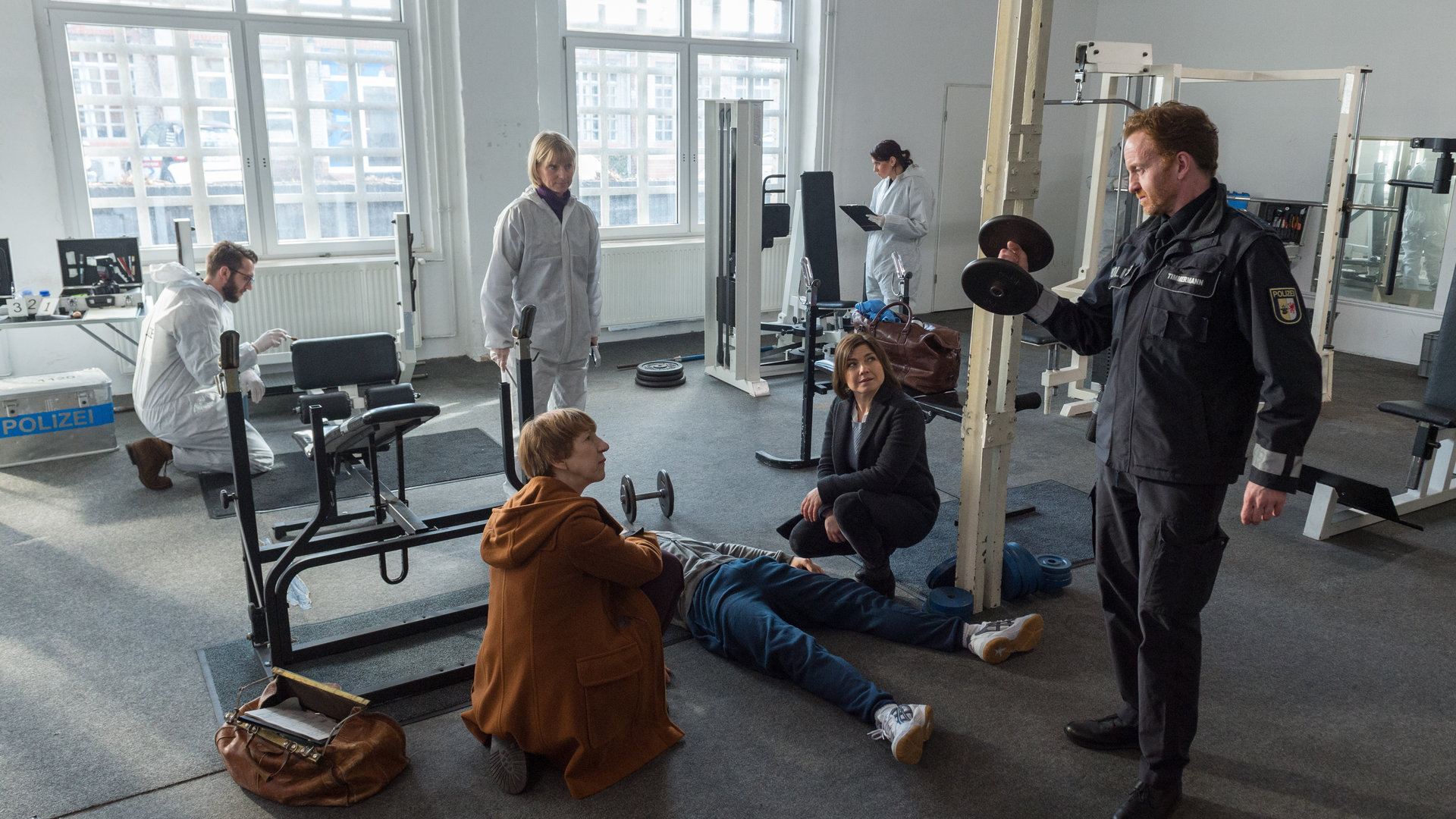 "SOKO Wismar – Knock-out": Zwischen Fitnessgeräten liegt die Leiche von Ronny Schnell (Mark Filatov). Daneben hocken Helene Sturbeck (Katharina Blaschke) und Katrin Börensen (Claudia Schmutzler). Sie schauen auf Timmermann (Mathias Junge), der eine Hantel hochhält. Im Hintergrund sind Roswitha Prinzler (Silke Matthias) und die KTU bei der Arbeit.