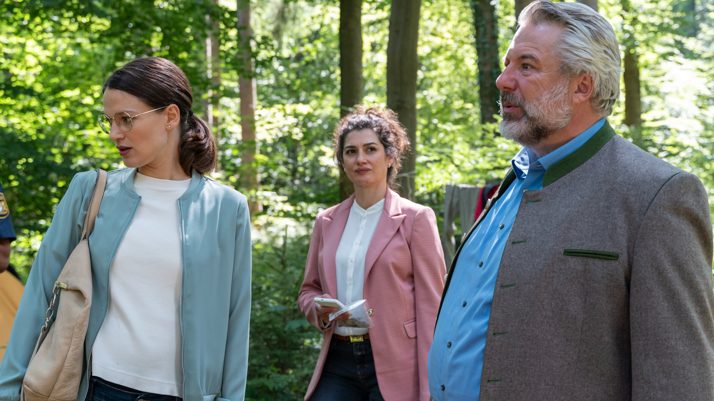 "Die Rosenheim-Cops - Eine Nacht im Wald": Kommissarin Beck (Michaela Weingartner), Rechtsmedizinerin Dimos (Anastasia Papadopoulou) und Kommissar Stadler (Dieter Fischer) stehen im Wald. Im Hintergrund steht ein Polizist (Komparse) in dunkelblauer Dienstkleidung.