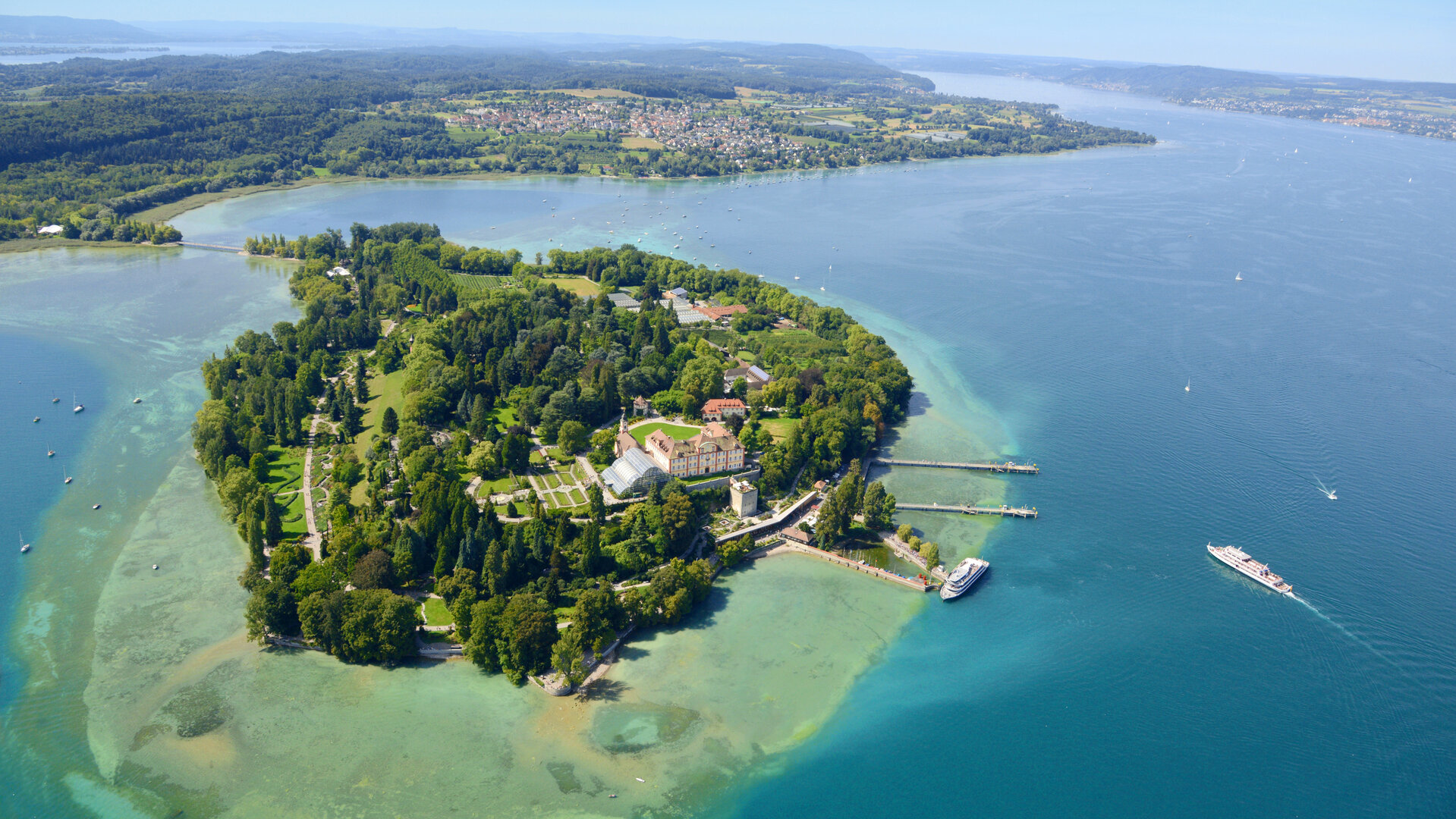 "Evangelischer Gottesdienst - Schöne Aussichten": Blick von schräg oben auf die sonnenverwöhnte Insel Mainau im Bodensee: Sie ist dicht bewaldet und von türkisblauem Wasser umgeben.