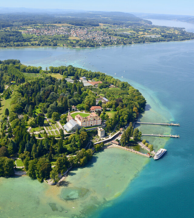 "Evangelischer Gottesdienst - Schöne Aussichten": Blick von schräg oben auf die sonnenverwöhnte Insel Mainau im Bodensee: Sie ist dicht bewaldet und von türkisblauem Wasser umgeben.