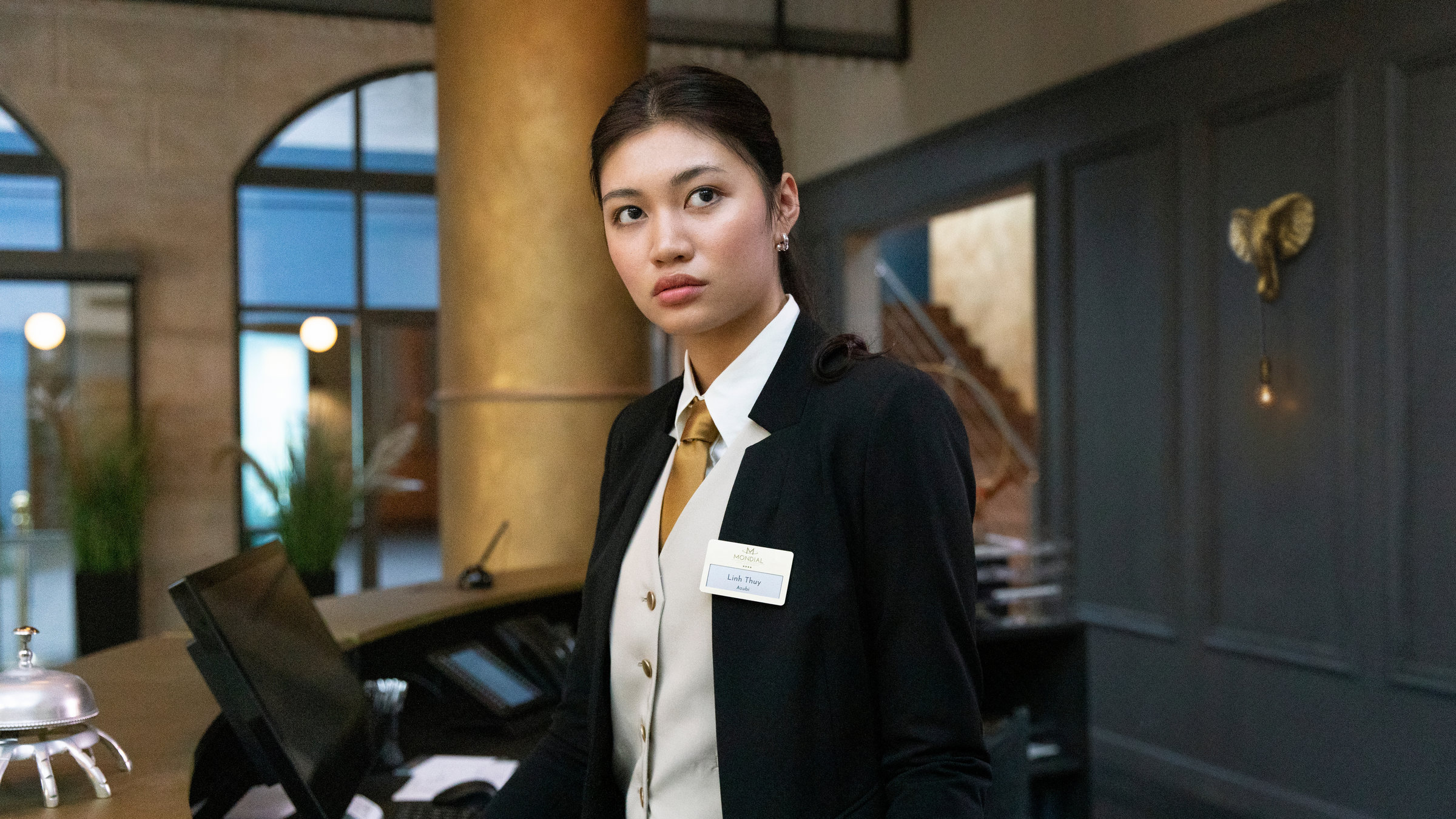 "Hotel Mondial - Zurück auf Los": Linh (Nhung Hong) steht hinter der Hotelrezeption und blickt in die Ferne. Auf dem Tresen der Rezeption stehen ein Bildschirm, eine Tastatur, ein Telefon und eine Klingel.