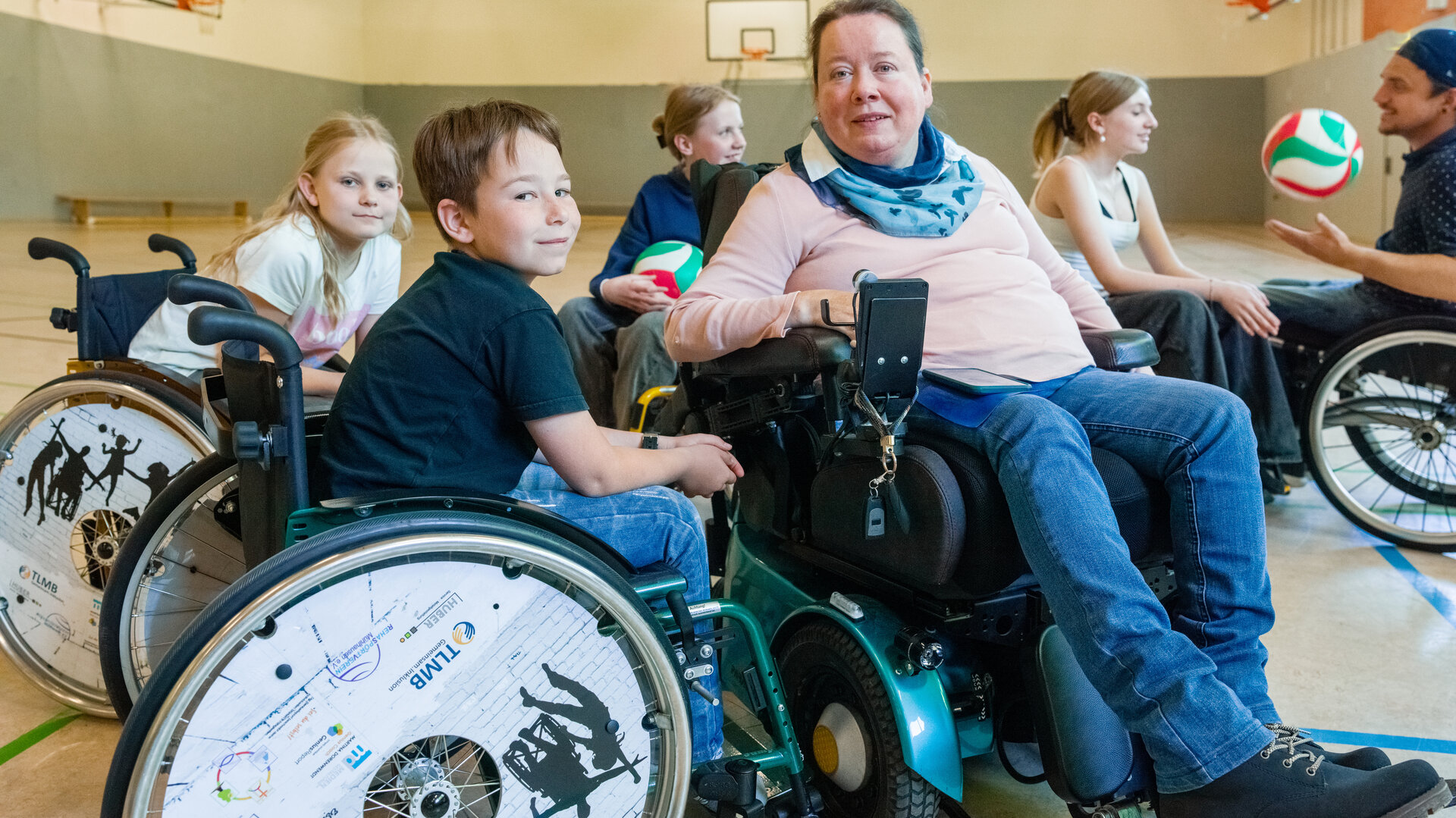 "einfach Mensch - Martina Dorenwendt: Ich lebe ohne Hindernisse": Martina Dorenwendt sitzt im Rollstuhl in einer Turnhalle. Um sie herum befinden sich einige Kinder in Rollstühlen, teilweise halten diese einen Ball.