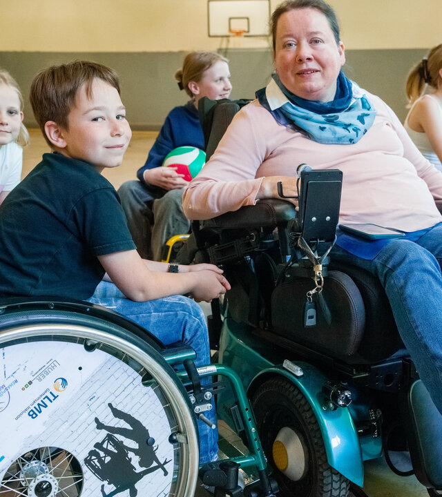 "einfach Mensch - Martina Dorenwendt: Ich lebe ohne Hindernisse": Martina Dorenwendt sitzt im Rollstuhl in einer Turnhalle. Um sie herum befinden sich einige Kinder in Rollstühlen, teilweise halten diese einen Ball.