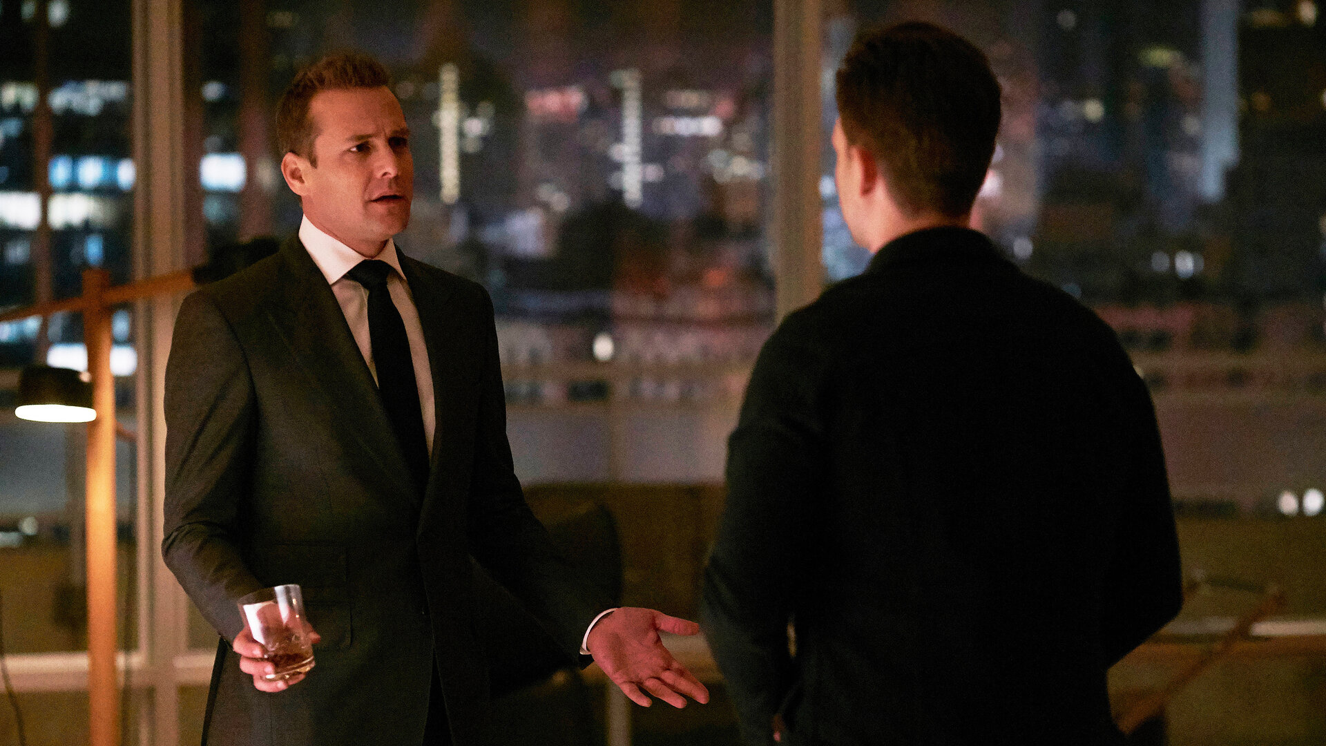 "Suits - Wem die Stunde schlägt": Harvey (Gabriel Macht) und Mike (Patrick J. Adams)  stehen abends im beleuchteten Büro und unterhalten sich. Harvey hält dabei einen Drink in seiner rechten Hand, während er gestikuliert.
