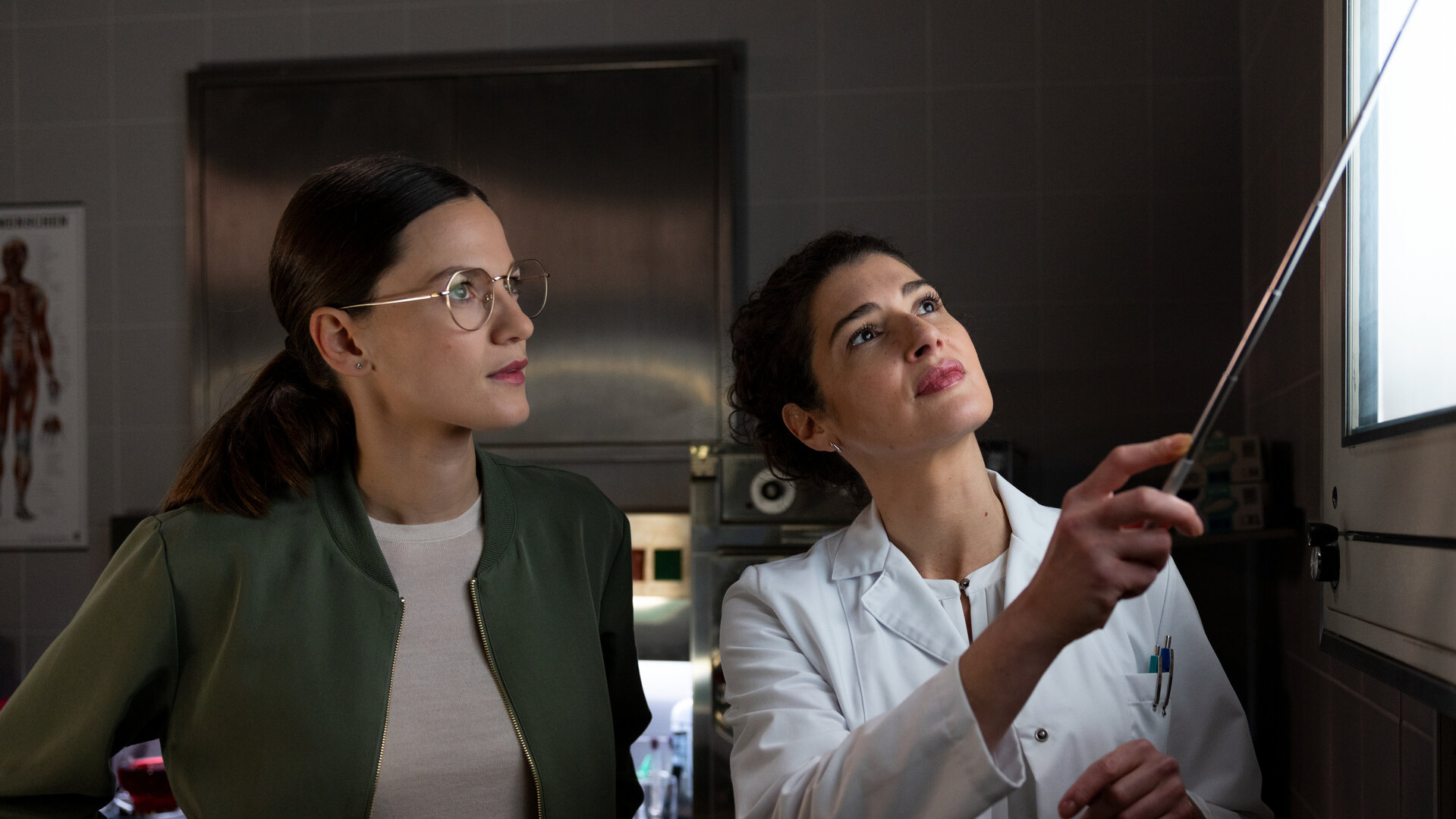 "Die Rosenheim-Cops - Das verflixte Paket": Hauptkommissarin Beck (Michaela Weingartner) und Rechtsmedizinerin Dr. Dimos (Anastasia Papadopoulou) stehen vor einem Leuchtkasten und betrachten ein Röntgenbild, auf das Dr. Dimos mit Hilfe eines Metallstabes zeigt.
