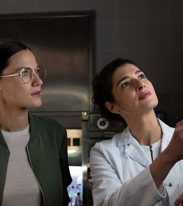 "Die Rosenheim-Cops - Das verflixte Paket": Hauptkommissarin Beck (Michaela Weingartner) und Rechtsmedizinerin Dr. Dimos (Anastasia Papadopoulou) stehen vor einem Leuchtkasten und betrachten ein Röntgenbild, auf das Dr. Dimos mit Hilfe eines Metallstabes zeigt.