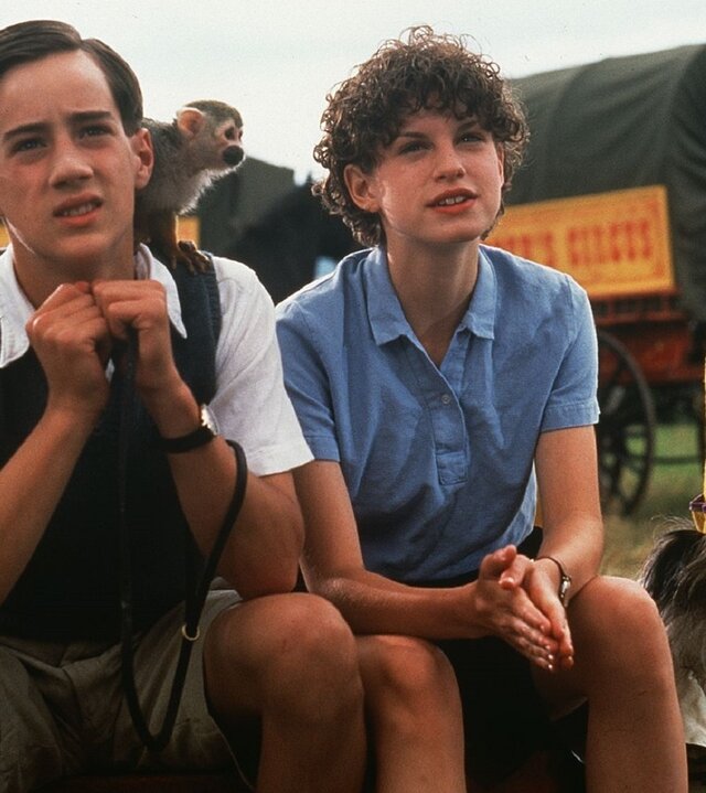 "Fünf Freunde und der Zauberer Wu": Tinker (James Tomlinson) und George (Jemima Rooper) sitzen nebeneinander auf einer Wiese. Tinker hat einen kleinen Affen auf der linken Schulter sitzen. Timmy der Hund sitzt links von George (Jemima Rooper) und trägt einen hohen gelb-lila Hut mit bunten Bommeln und Rüschen um seinen Hals. Hinter den vier sind zwei Kutschwägen mit Schildern zu sehen. Daneben steht ein kostümierter Mann mit jeweils einer brennenden Fackel in der Hand.