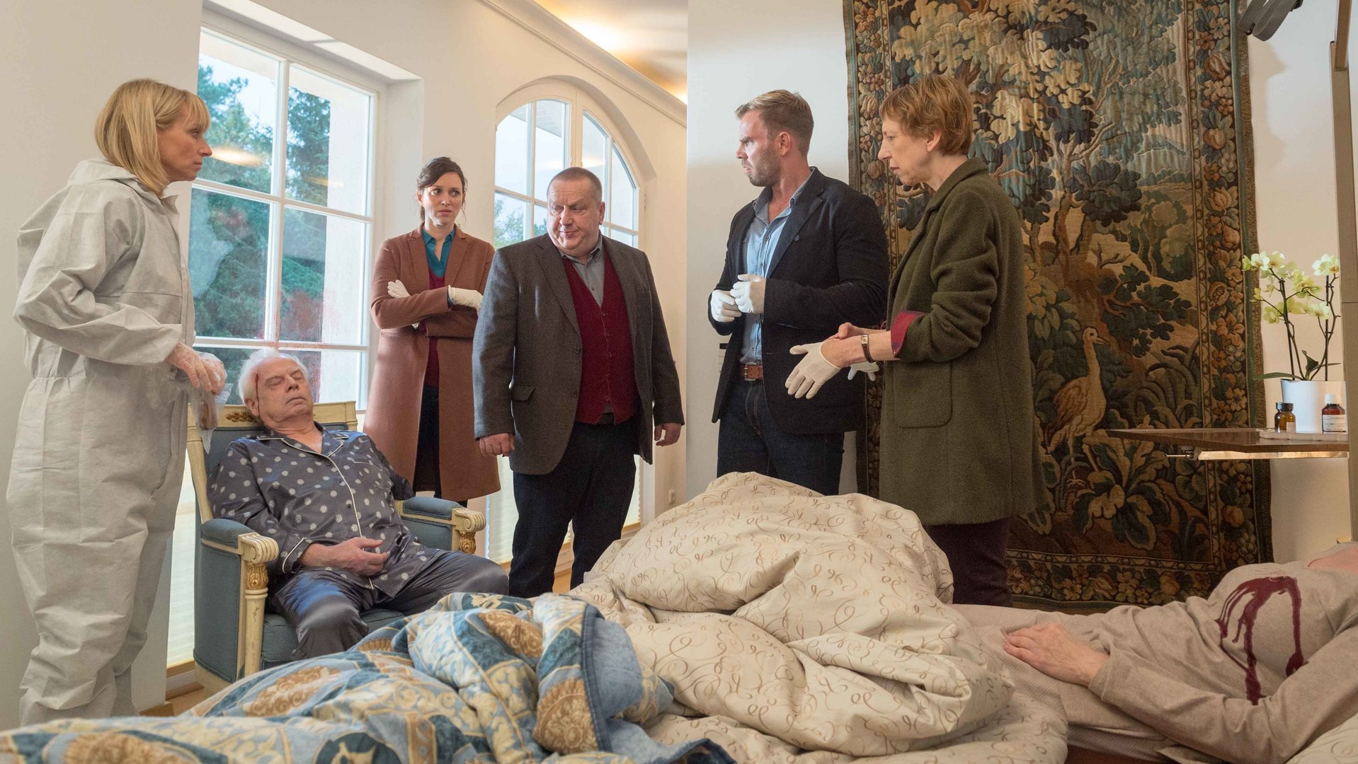 "SOKO Wismar – Alte Freunde": Wilhelm Kusche (Heiner Niedung) sitzt eingesackt im Sessel. Gegenüber im Bett liegt Rosa Kusche (Beate Häbich-Cardoso Dos Santos) mit Wunde am Herzen. Ringsum stehen Rechtsmedizinerin Helene Sturbeck (Katharina Blaschke) und die Ermittler Joost (Nike Fuhrmann), Reuter (Udo Kroschwald) und Pöhlmann (Dominic Boeer) sowie Kriminaltechnikerin Roswitha Prinzler (Silke Matthias).