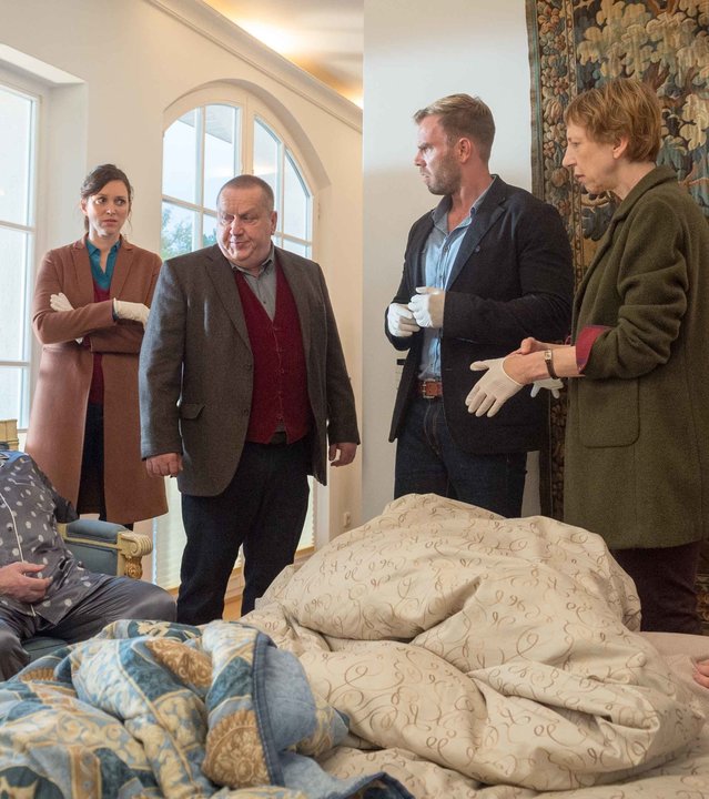 "SOKO Wismar – Alte Freunde": Wilhelm Kusche (Heiner Niedung) sitzt eingesackt im Sessel. Gegenüber im Bett liegt Rosa Kusche (Beate Häbich-Cardoso Dos Santos) mit Wunde am Herzen. Ringsum stehen Rechtsmedizinerin Helene Sturbeck (Katharina Blaschke) und die Ermittler Joost (Nike Fuhrmann), Reuter (Udo Kroschwald) und Pöhlmann (Dominic Boeer) sowie Kriminaltechnikerin Roswitha Prinzler (Silke Matthias).