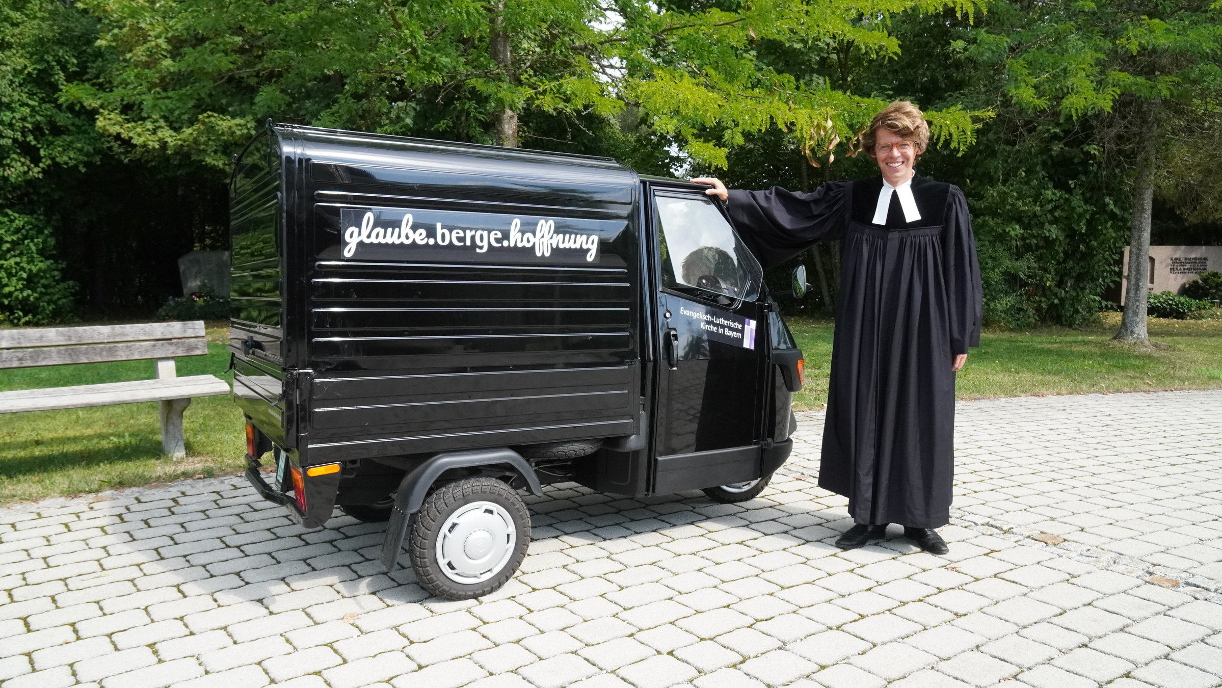 "37°: Kirche mal anders - Pastor*innen im Aufbruch": Ein schwarzes Kleintransporter-Dreirad italienischer Bauart steht von links nach rechts gerichtet auf einem gepflasterten Platz. An seiner Seite prangt der Schriftzug "Glaube. Berge. Hoffnung". Rechts davon steht der junge Penzberger Pastor Philipp Roß in schwarzem Talar mit weißem Beffchen, blickt in die Kamera und stützt seine rechte Hand auf dem Fahrzeug ab. Den Hintergrund bestimmen grüne Bäume und Büsche.