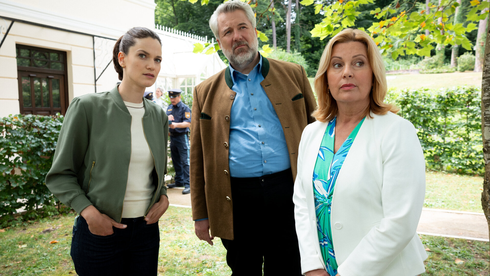 "Die Rosenheim-Cops - Zwei Koffer aus Berlin": Hauptkommissarin Beck (Michaela Weingartner), Hauptkommissar Stadler (Dieter Fischer) und Marie Hofer (Karin Thaler) stehen auf einem Rasen neben einem Baum und schauen in dieselbe Richtung. Im Hintergrund steht ein Haus vor Bäumen.