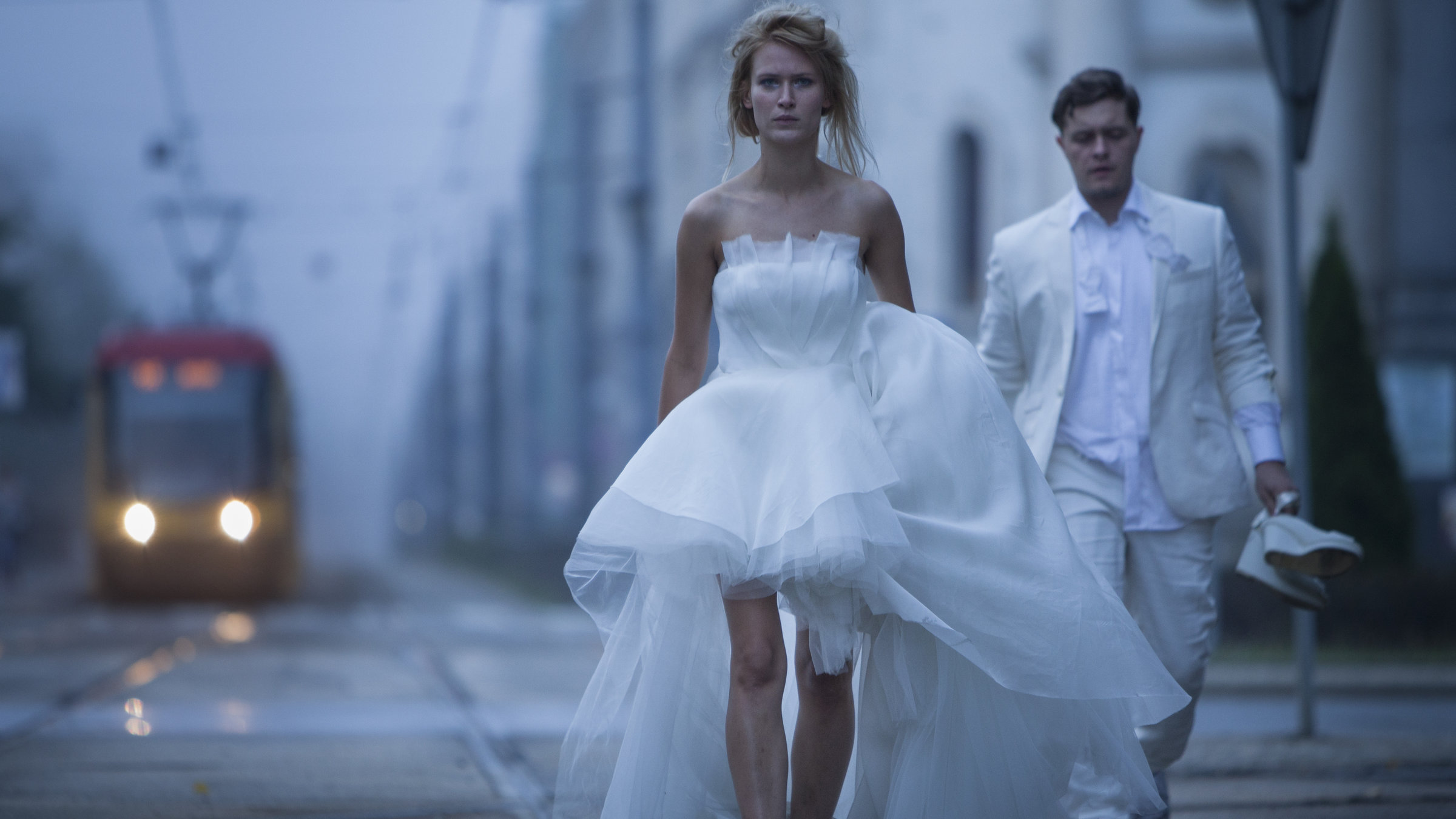 "Chemo": Lena (Agnieszka Żulewska) im langen weißen Brautkleid geht mit ausdruckslosem Gesicht und aufgelösten Haaren im Morgengrauen barfuß auf einer menschenleeren Großstadtstraße. Links neben ihr fährt eine Straßenbahn heran, rechts trägt ihr Benek (Tomasz Schuchardt)  im weißen Hochzeitsanzug die Schuhe hinterher.