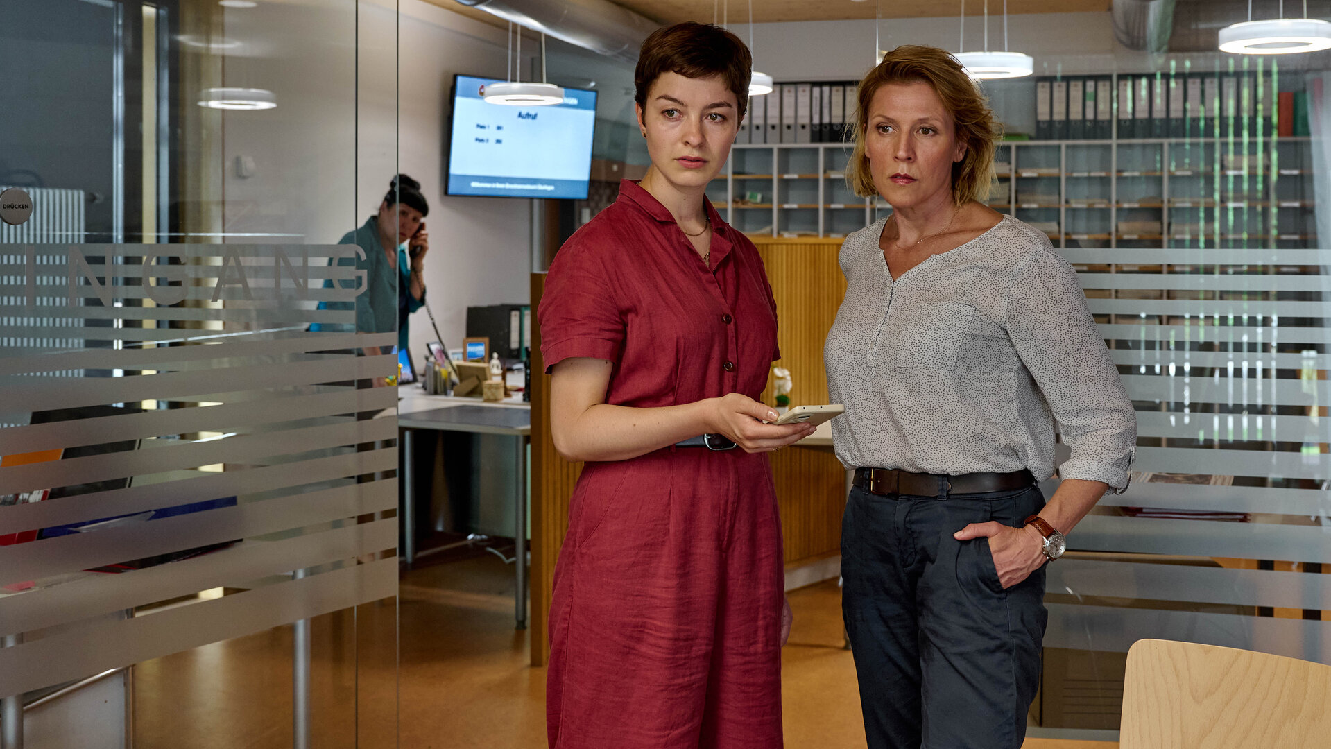 "Mordufer - Die große Gier": Chiara Locatelli (Maria Wördemann) und Doro Beitinger (Franziska Weisz) stehen in einem Büro und blicken zur Seite. Locatelli hält ein Smartphone in ihrer rechten Hand. Im Hintergrund steht Katrin Glaunsinger (Beate Constanze Pitronik) an einem Schreibtisch und telefoniert mit einem Festnetztelefon.