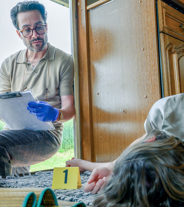 "SOKO Linz - Herzstiche": Gerichtsmediziner Dr. Richard "Richie" Vitek (Alexander Pschill) lehnt sich in eine offene Tür von einem Camper, durch die er zu einer Leiche von Katharina Hofmeister (Valentina Inzko Fink) schaut, die auf dem Boden liegt. Er trägt Schutzhandschuhe und protokolliert.