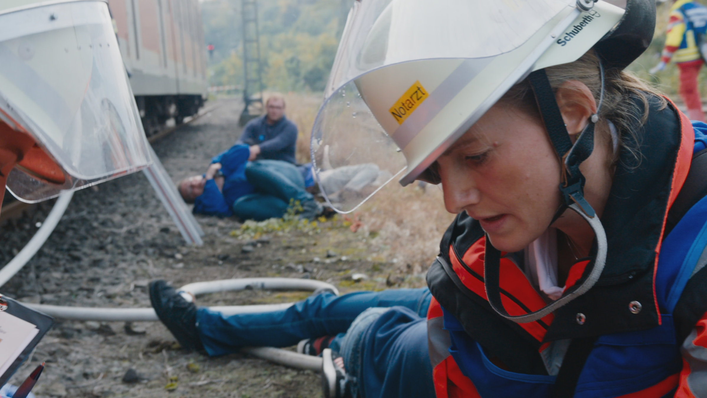Junge blonde Frau hilft einem Verletzten. Im Hintergrund suchen zwei Männer nach weiteren Verletzten.
