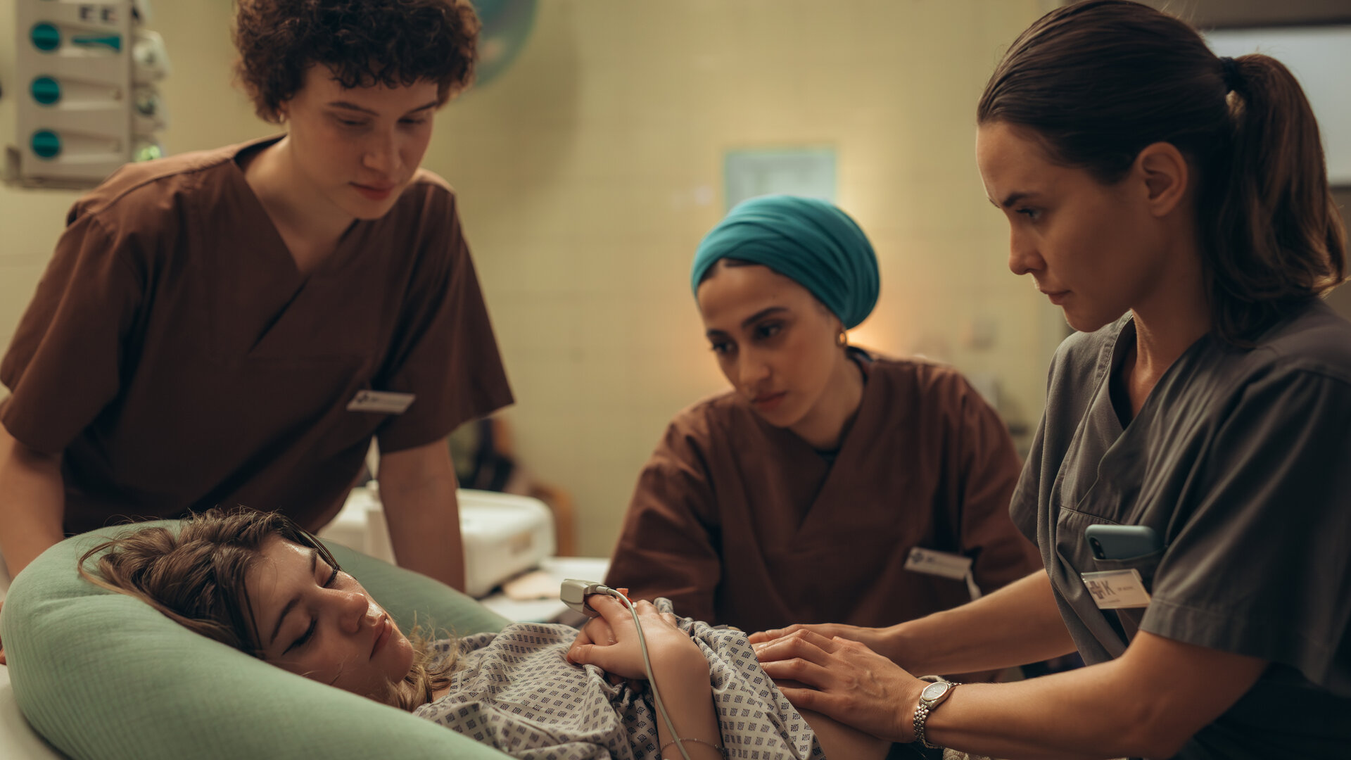 "PUSH - Geheimnisse": Lou (Lisa Moell) liegt in der Klinik. Greta (Lydia Amasko) und Meryem (Tua El-Fawwal) stehen daneben während Dr. Mohn (Katia Fellin) Lou untersucht.