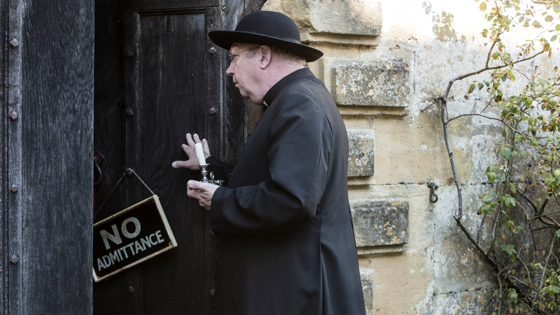"Father Brown - Abschied von Lady Felicia": Father Brown (Mark Williams) hält mit einer Hand eine Holztür geöffnet, in der anderen hält er eine Kerze. An der Tür hängt ein Schild mit der Aufschrift ''No Admittance''.