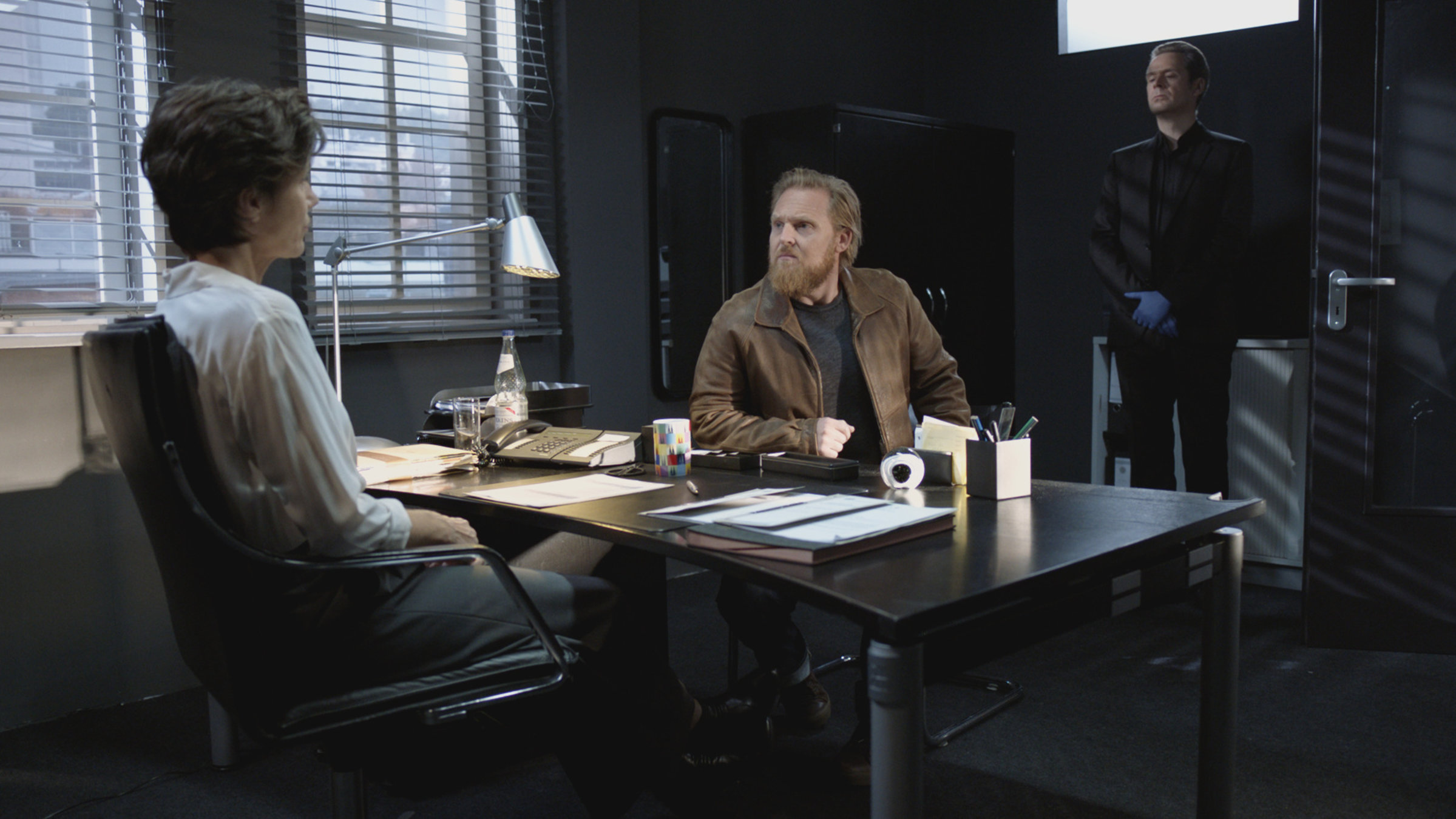 "Professor T. - Rache": Christina Fehrmann (Julia Bremermann) sitzt mit dem Rücken zur Kamera an ihrem Schreibtisch. Ihr gegenüber am Tisch sitzt Thomas Reuter (Axel Stein), der seinen Arm auf den Tisch gestützt hat. Im Hintergrund steht Professor T. (Matthias Matschke), der das Gespräch der beiden verfolgt.