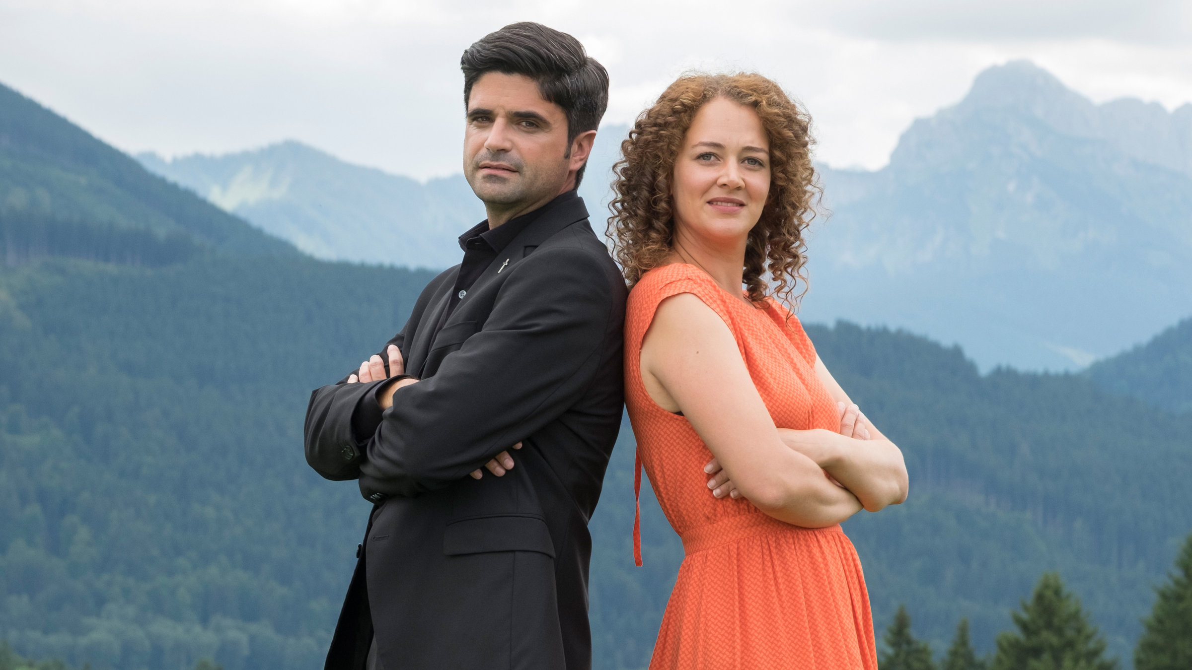 "Tonio & Julia - Kneifen gilt nicht": Mit dem Rücken aneinandergelehnt und nach vorne in die Kamera blickend, stehen Pfarrer Tonio Niederegger (Maximilian Grill) und Familientherapeutin Julia Schindel (Oona Devi Liebich) auf einer Wiese. Im Bildhintergrund Bäume und Berge.