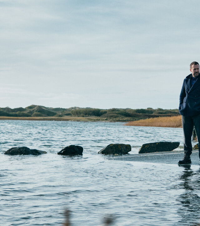 "Under Salt Marsh - Tod in den Salzwiesen - Das GestÃ¤ndnis": Eric Bull (Rafe Spall) und Jess Deng (Naomi Yang) stehen im Freien auf einer StraÃe, die zum Teil Ã¼berflutet ist. Seitlich von ihnen steht ein groÃes Warnschild.